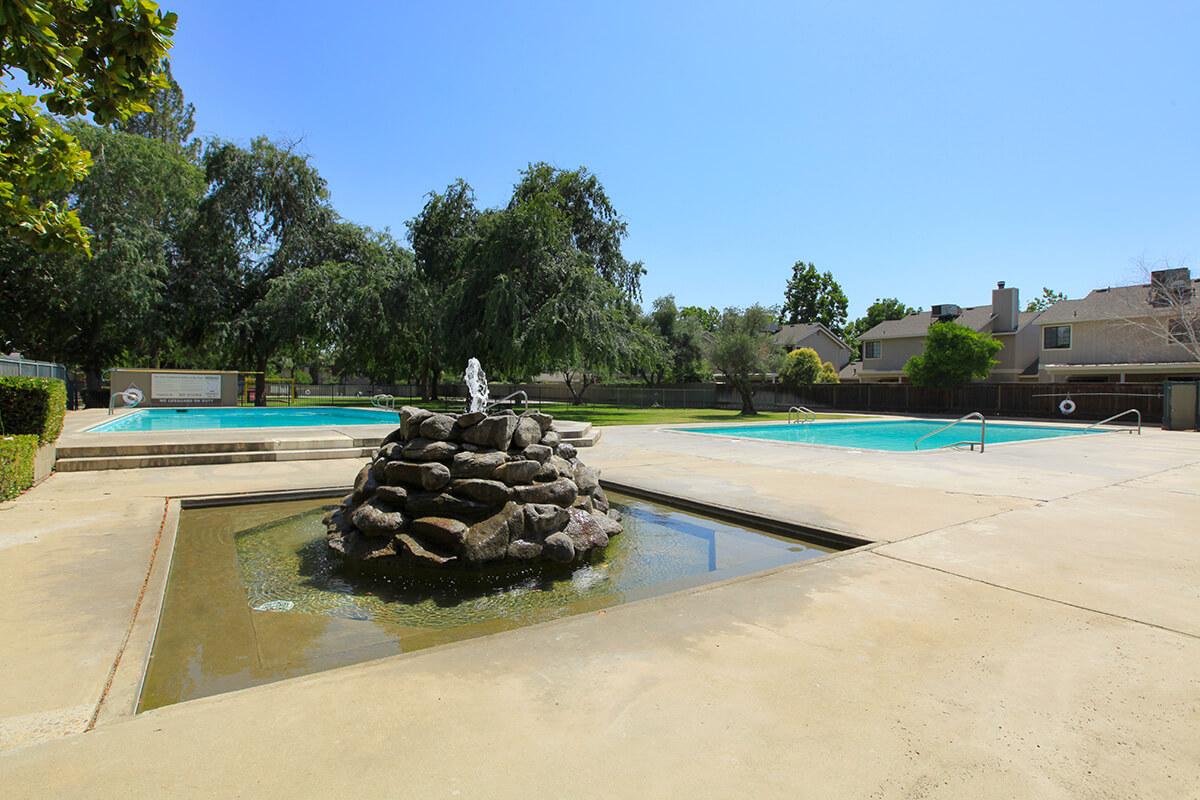 A sunny pool area featuring two swimming pools, a rock fountain in the foreground, and lush greenery surrounding the space. The sky is clear blue, and there are a few trees and residential buildings in the background.