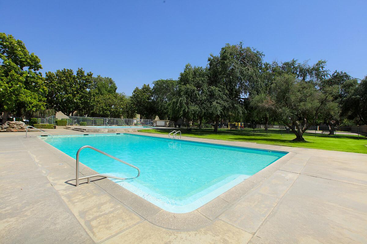 A clear blue swimming pool surrounded by green grass and trees. The sun is shining in a clear blue sky, creating a bright and inviting atmosphere. There are safety rails on the edge of the pool, and a fenced area in the background with more trees. Ideal for relaxation and outdoor activities.