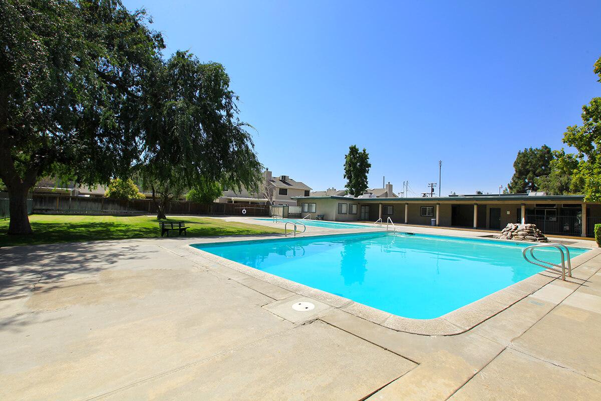 A clear blue swimming pool surrounded by a concrete patio. Lush green trees and a grassy area are visible nearby. Several buildings are in the background under a bright blue sky. The setting appears peaceful and inviting, suitable for relaxation.