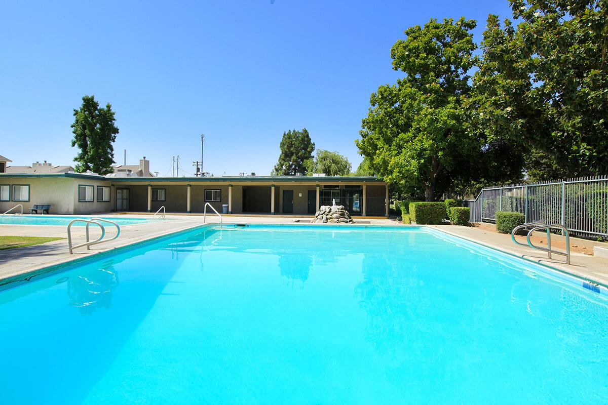 A clear swimming pool surrounded by a fence, with lounge chairs nearby. Green trees and a building are visible in the background, under a bright blue sky. The water is calm and inviting, reflecting the sunlight.