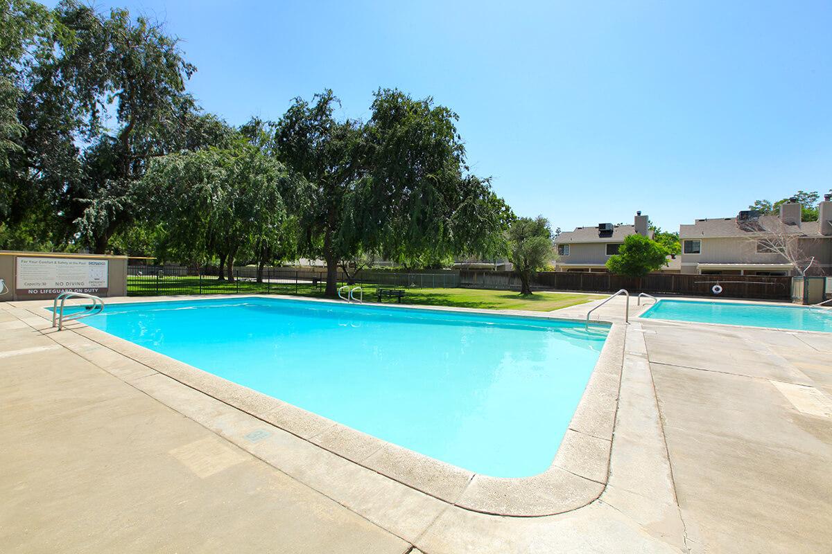 A bright outdoor swimming pool with clear blue water, surrounded by green grass and trees. In the background, there are residential buildings. The pool area features a concrete deck and a sign near the edge. The scene is set under a clear blue sky.