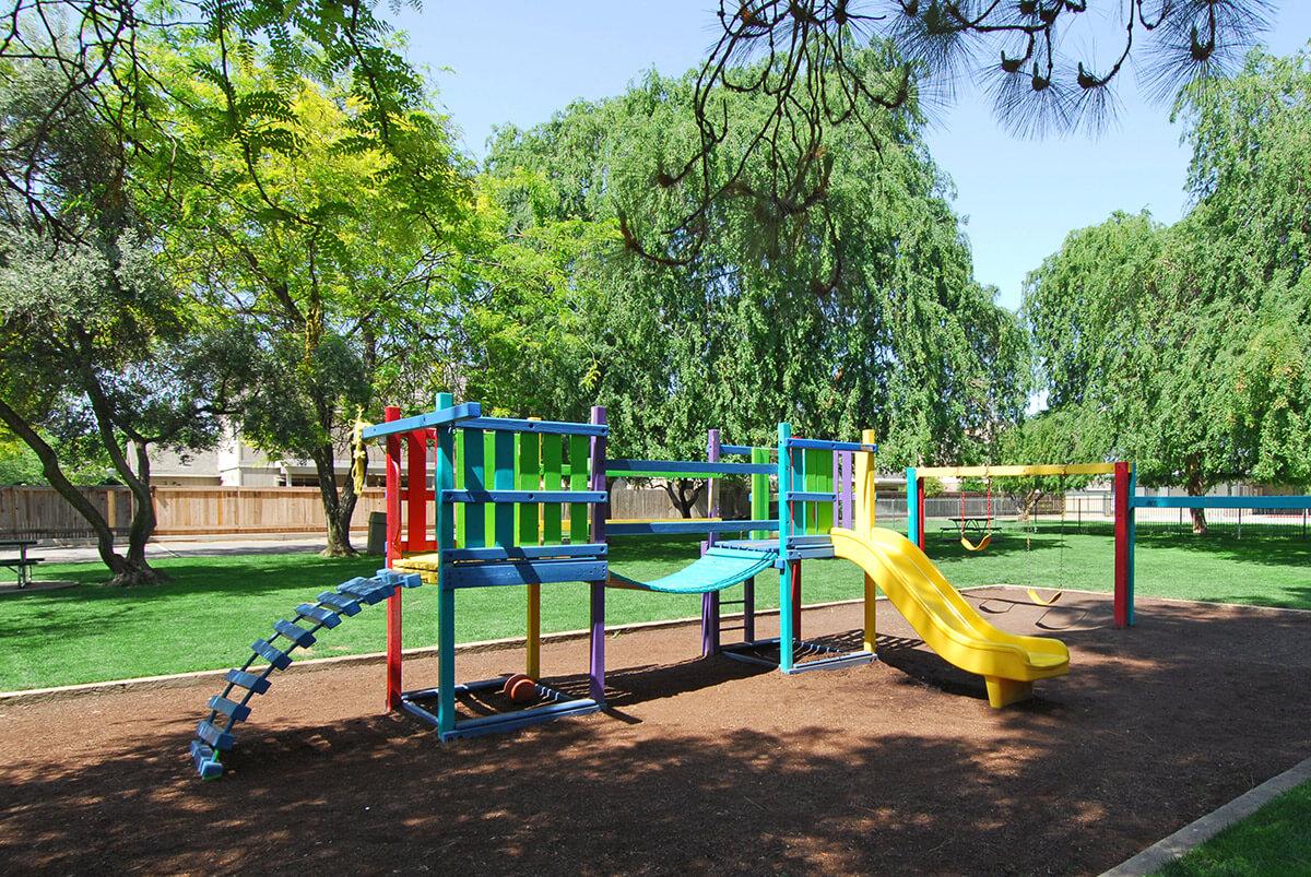 Colorful playground structure featuring a slide, climbing elements, and swings, set in a grassy area with trees. The scene is bright and sunny, creating a cheerful outdoor environment for children to play.