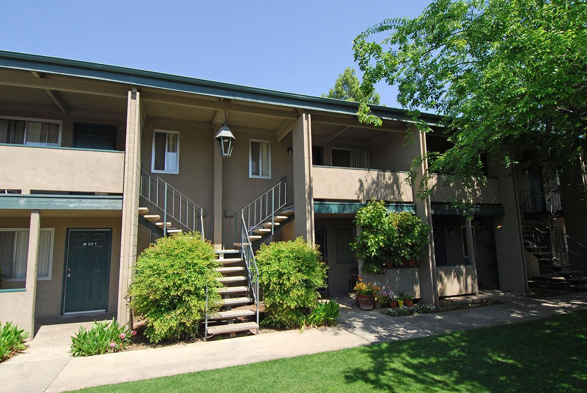 A two-story apartment building with a green roof and exterior staircases. The front features several potted plants and shrubs, with a clear blue sky above. The entrance doors are visible, and there are balconies on the upper floor. The setting appears well-maintained and inviting.