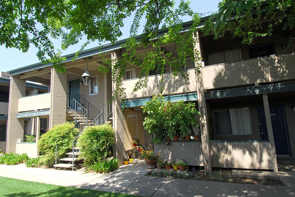 A two-story apartment building featuring a central staircase leading to the upper level. The building has balconies with potted plants and is surrounded by lush greenery. The ground level has a pathway and various shrubs, enhancing the inviting atmosphere of the community.