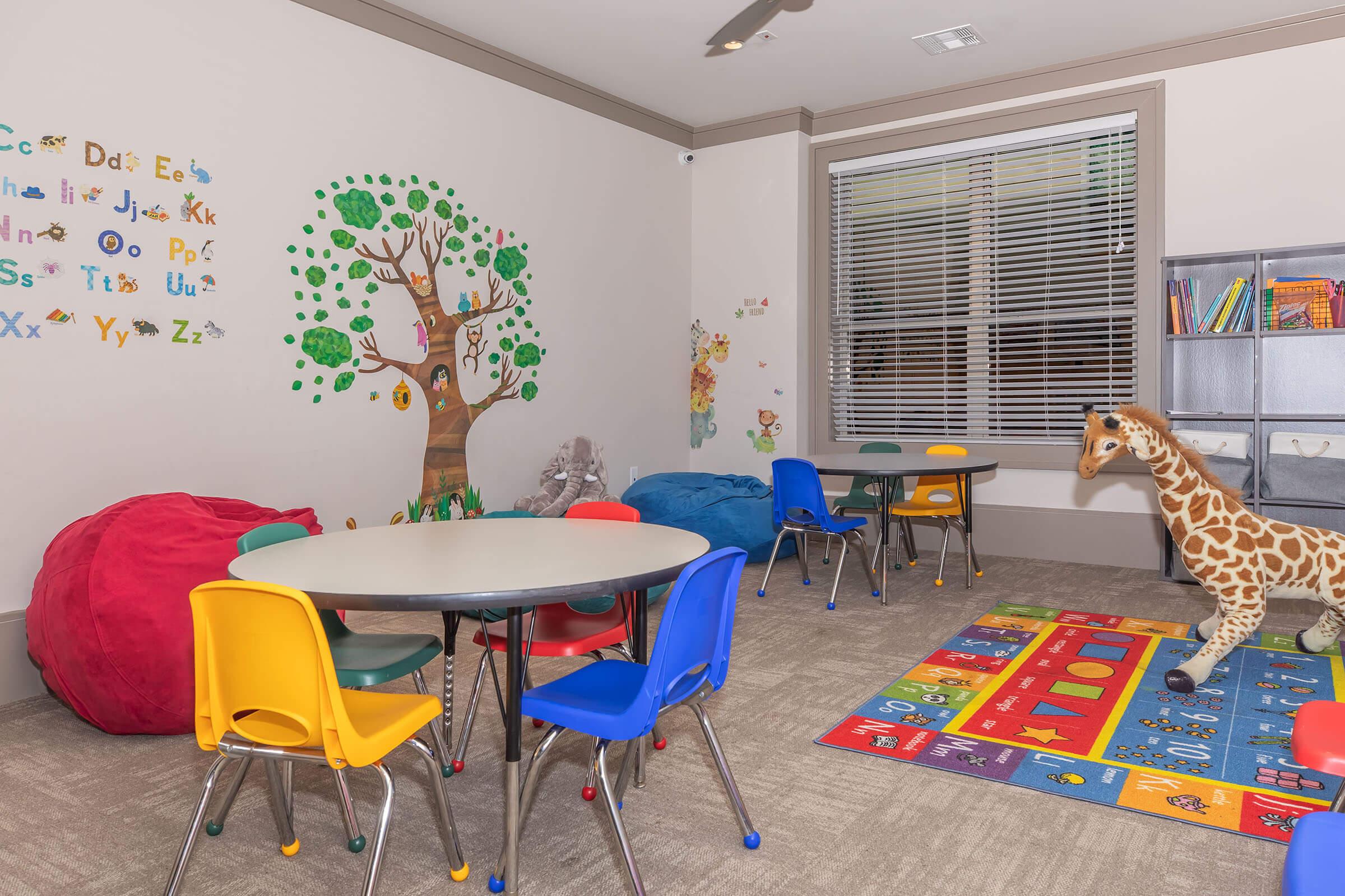 A colorful children's classroom with a tree mural on one wall, featuring playful letters and numbers. There are two tables with circular tops surrounded by red, blue, and yellow chairs. A vibrant area rug displays letters and numbers, while a stuffed giraffe adds a whimsical touch. Two large bean bags are also present.