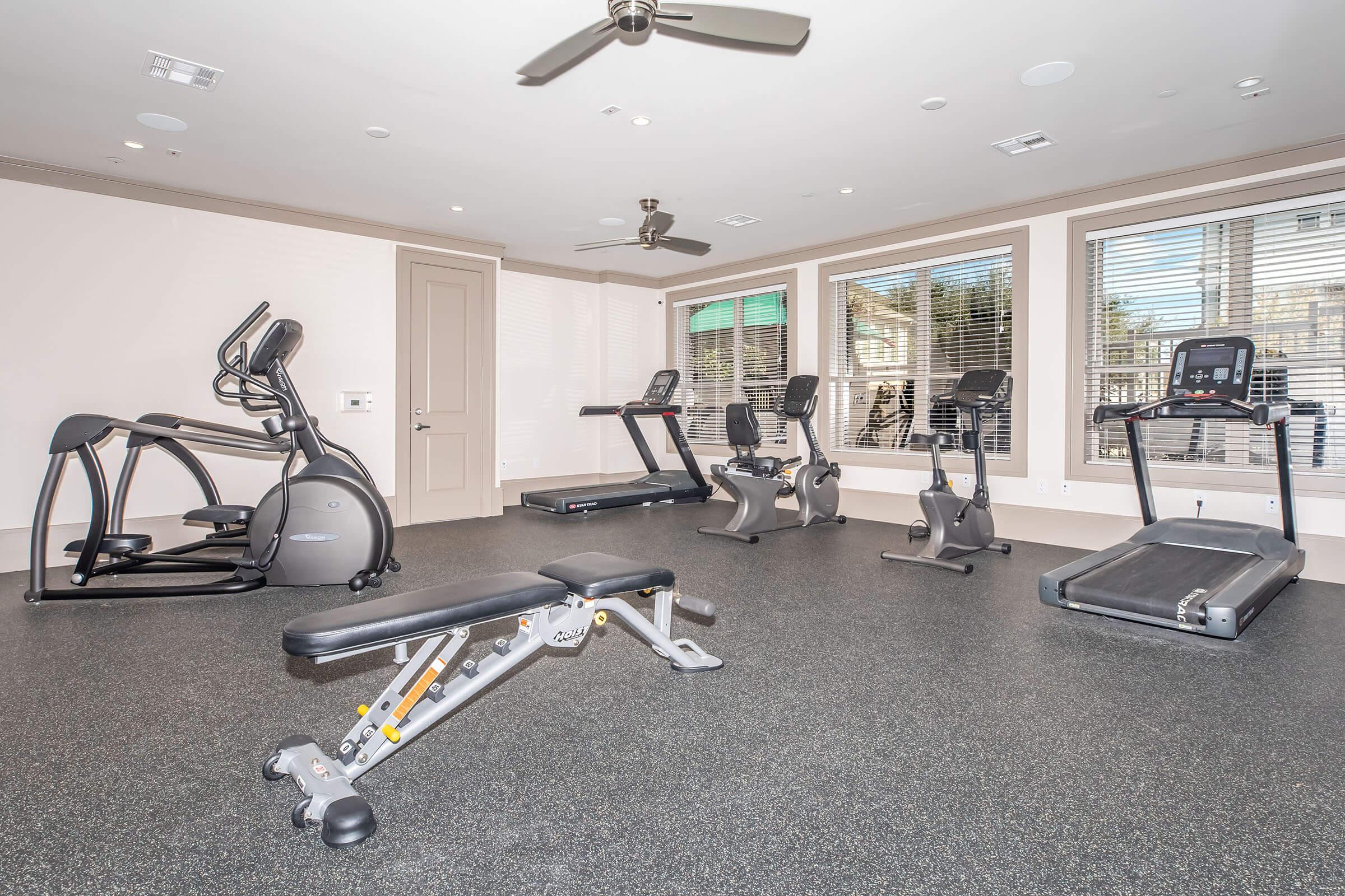 A modern gym interior featuring various fitness equipment, including an elliptical machine, stationary bike, treadmill, and weight bench. The room has large windows allowing natural light, and the flooring is covered with black rubber mats. Ceiling fans provide ventilation.