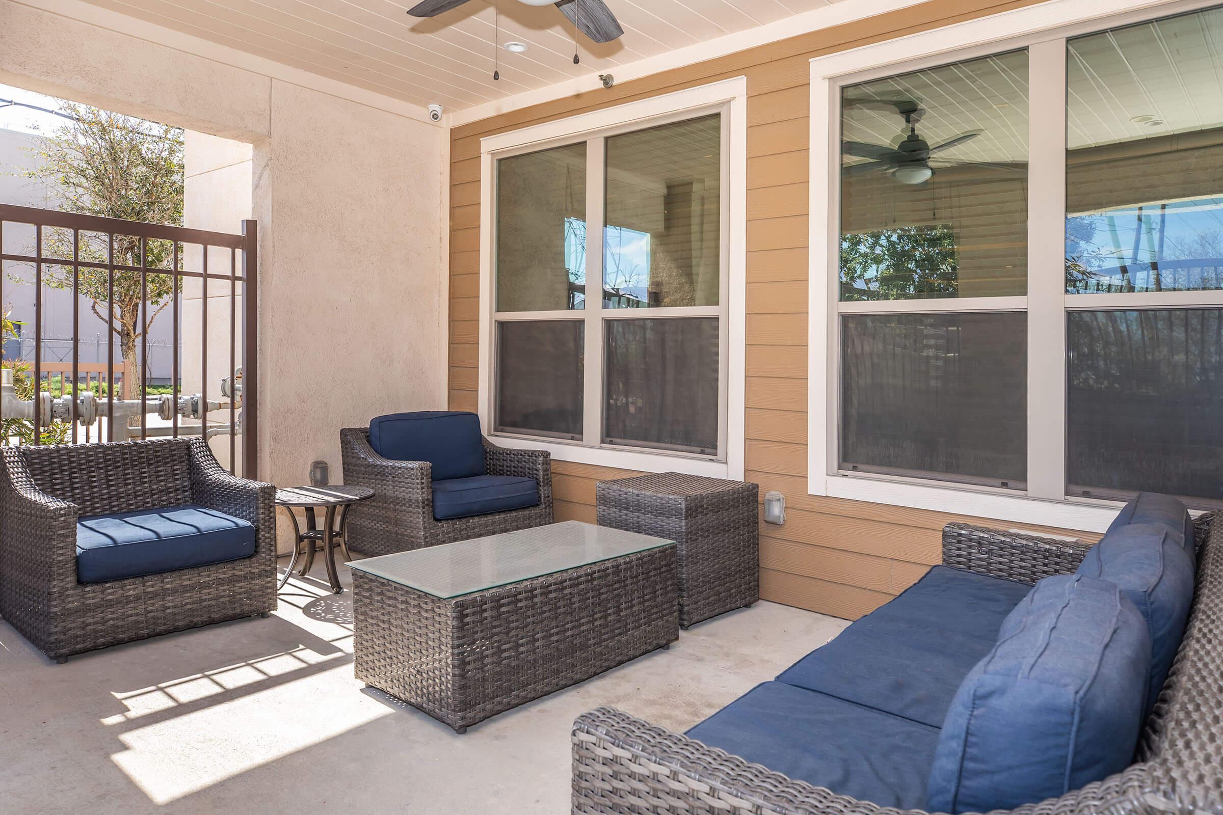 A cozy outdoor seating area featuring a glass coffee table, two armchairs, and a sofa with blue cushions, all made of woven rattan. The space is enclosed by a gate and has large windows allowing natural light. A fan is visible on the ceiling, enhancing the atmosphere for relaxation.