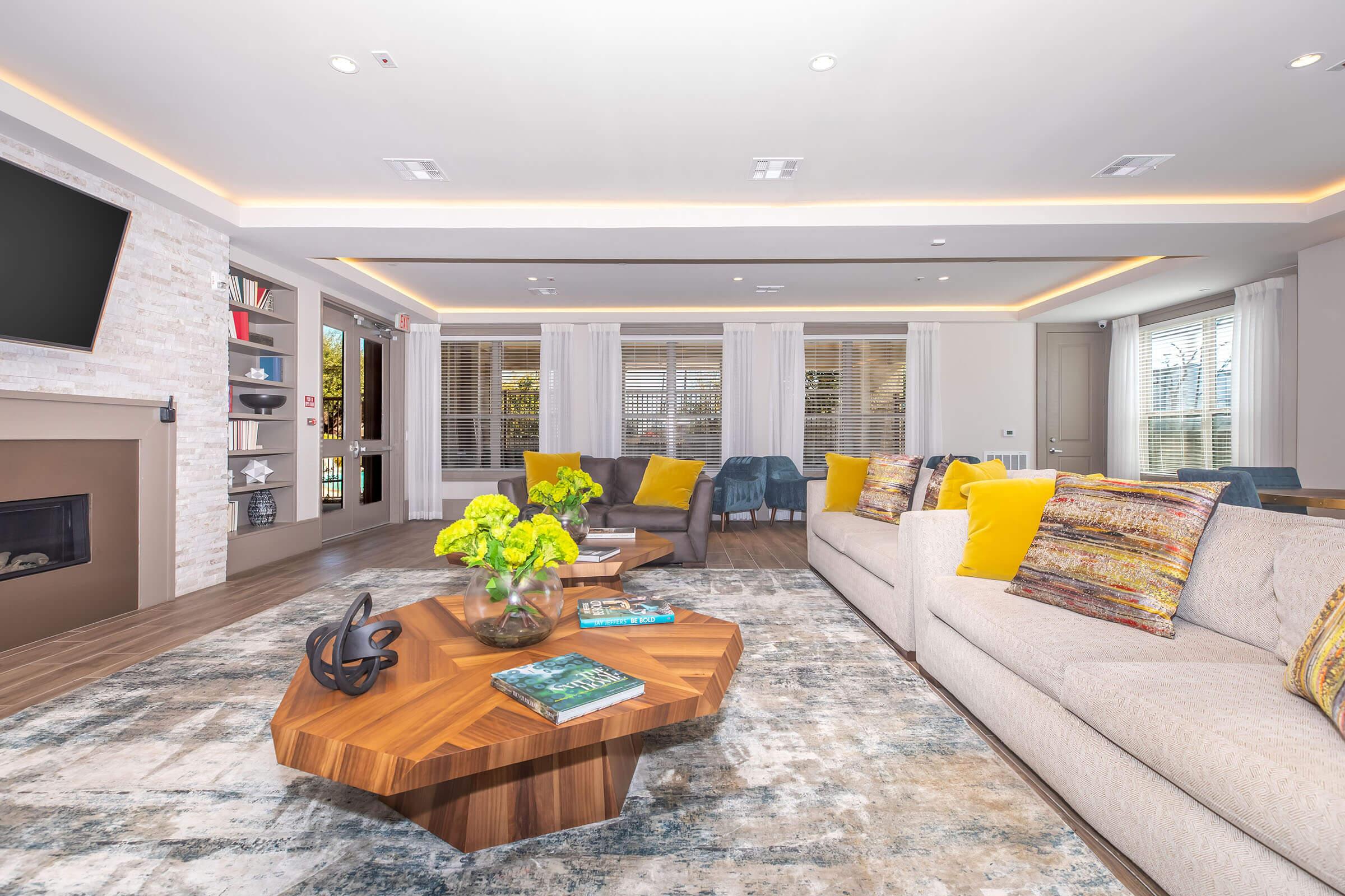 A modern living room featuring a cozy sofa with colorful throw pillows, a stylish coffee table with books and a decorative bowl, and a plush area rug. Large windows allow natural light, complemented by warm ceiling lights. Decor includes vibrant floral arrangements and contemporary art on the walls.