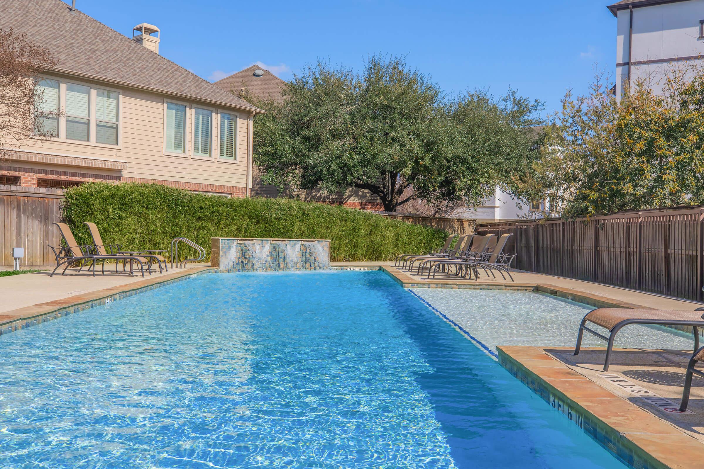 A clear blue swimming pool with a water feature, surrounded by lounge chairs. Green shrubs and trees line the pool area, with a wooden fence in the background. Sunlight casts bright reflections on the water, and a clear blue sky is visible overhead.