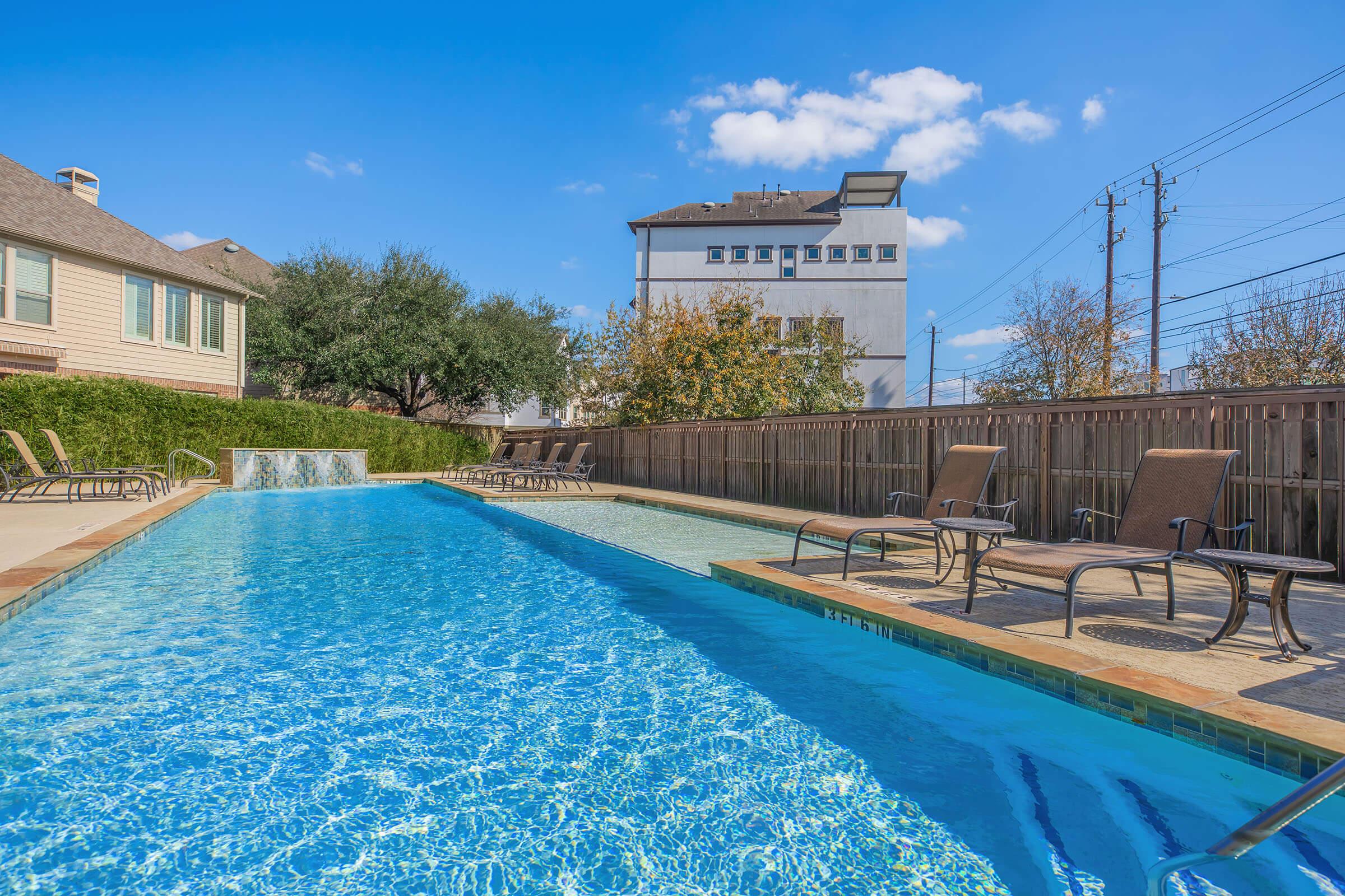 A bright, sunny outdoor pool area featuring a long, clear blue swimming pool with a waterfall feature. Surrounding the pool are brown lounge chairs and a wooden fence. In the background, there are several residential buildings and trees under a clear blue sky with a few scattered clouds.