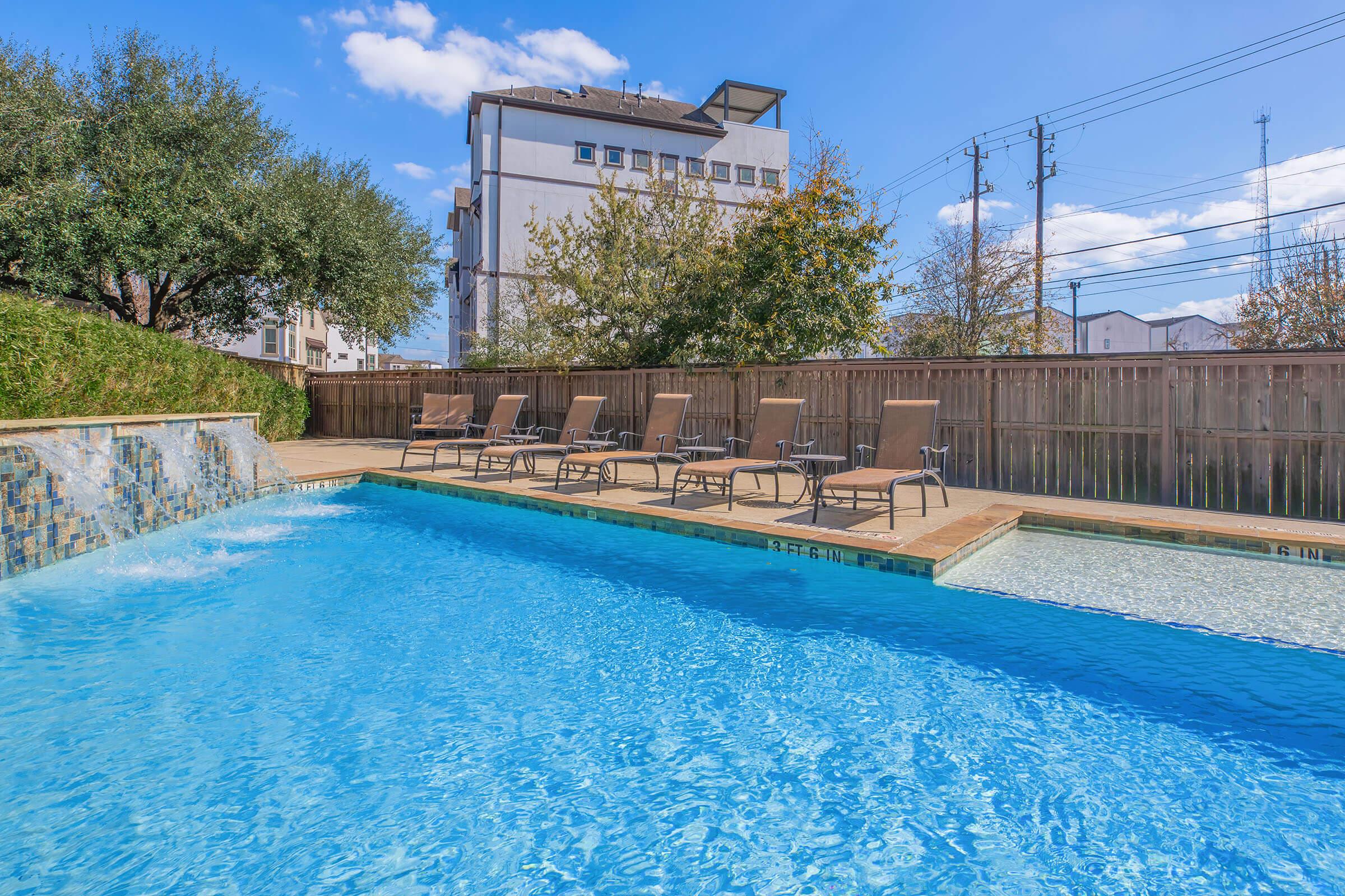 A serene swimming pool with crystal-clear water, featuring a decorative waterfall on one side. Sun loungers are neatly arranged along the edge of the pool, surrounded by lush greenery. In the background, a modern building and power lines are visible under a bright blue sky with fluffy clouds.