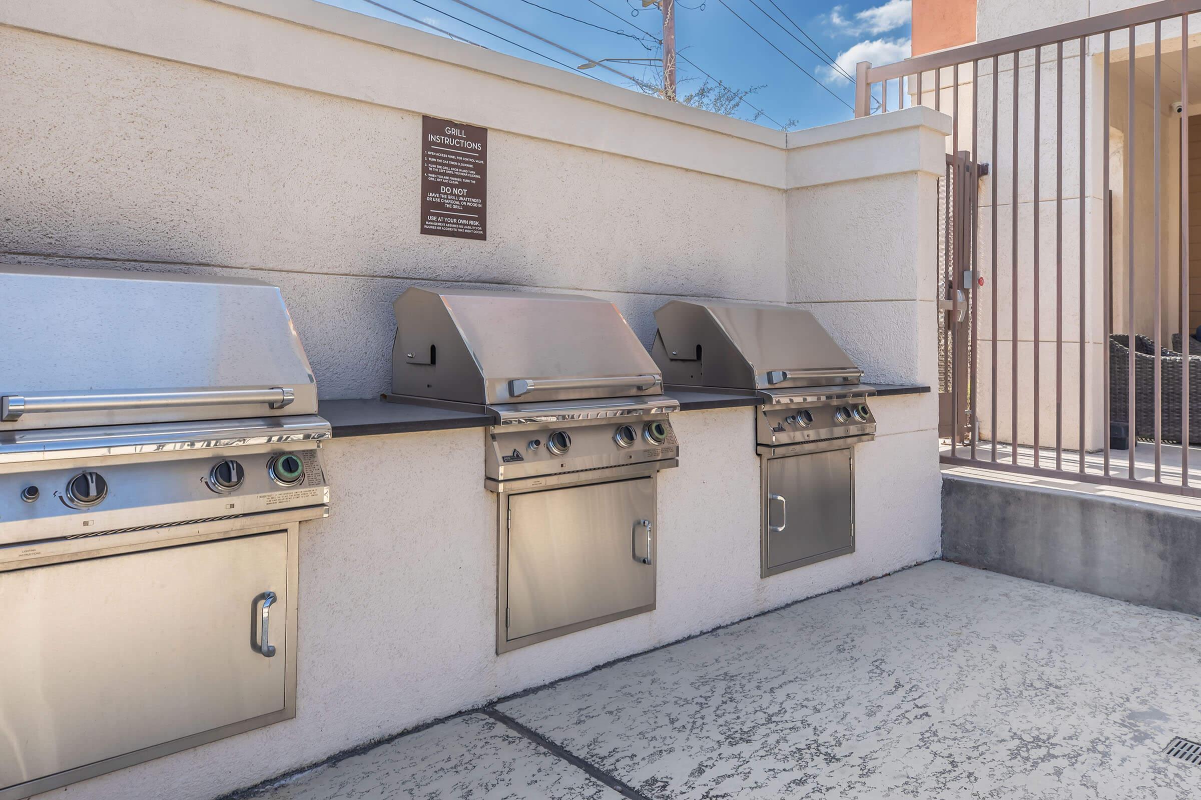 Three stainless steel outdoor barbecue grills situated against a light-colored wall. A sign is mounted above the grills, and a gated area is visible in the background, with blue skies and power lines overhead. The ground is paved with textured concrete.