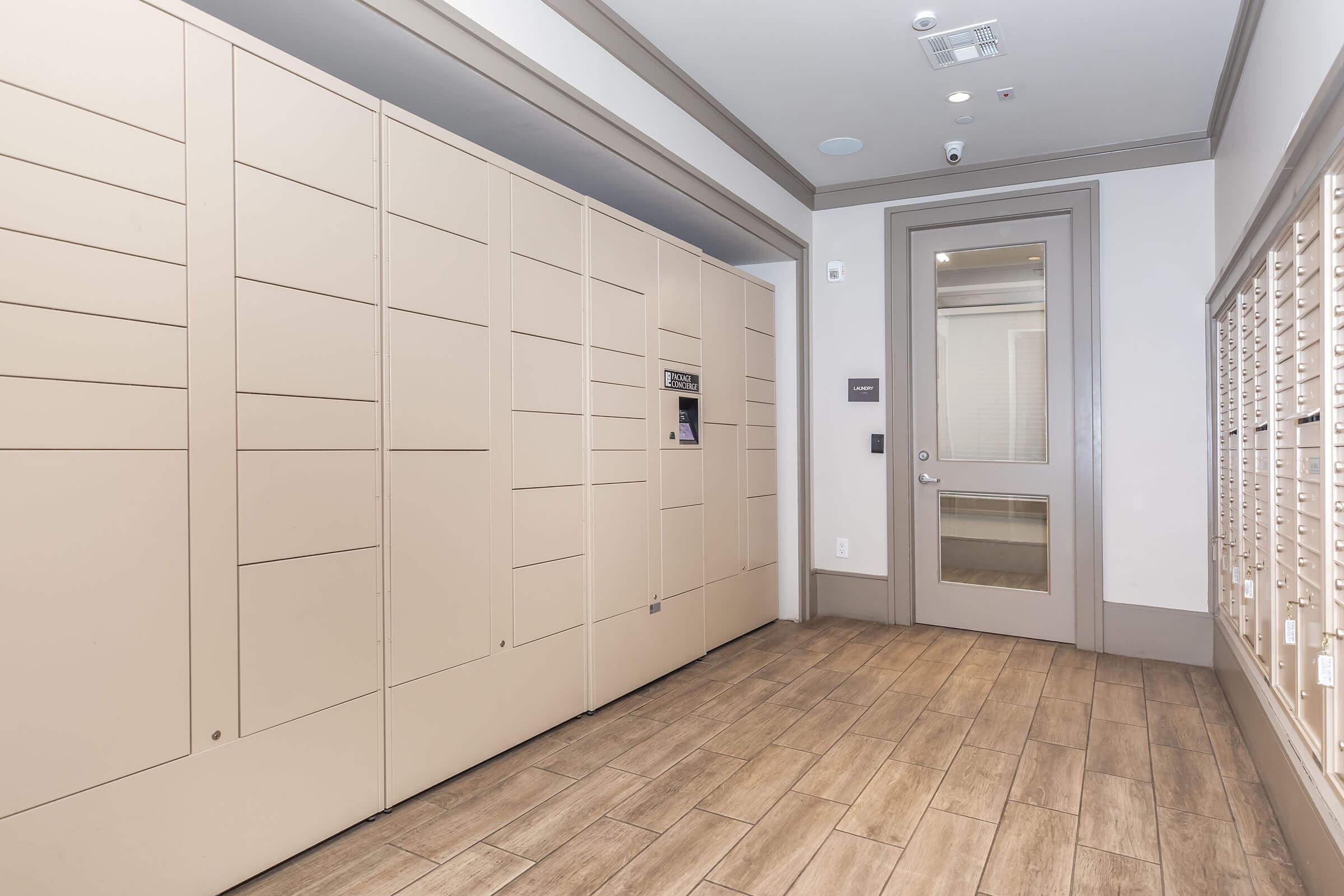 A modern parcel locker area featuring beige lockers against a wall, with a glass door leading to another room. The floor is covered in wooden tiles, and there are overhead lights and a small security camera visible.