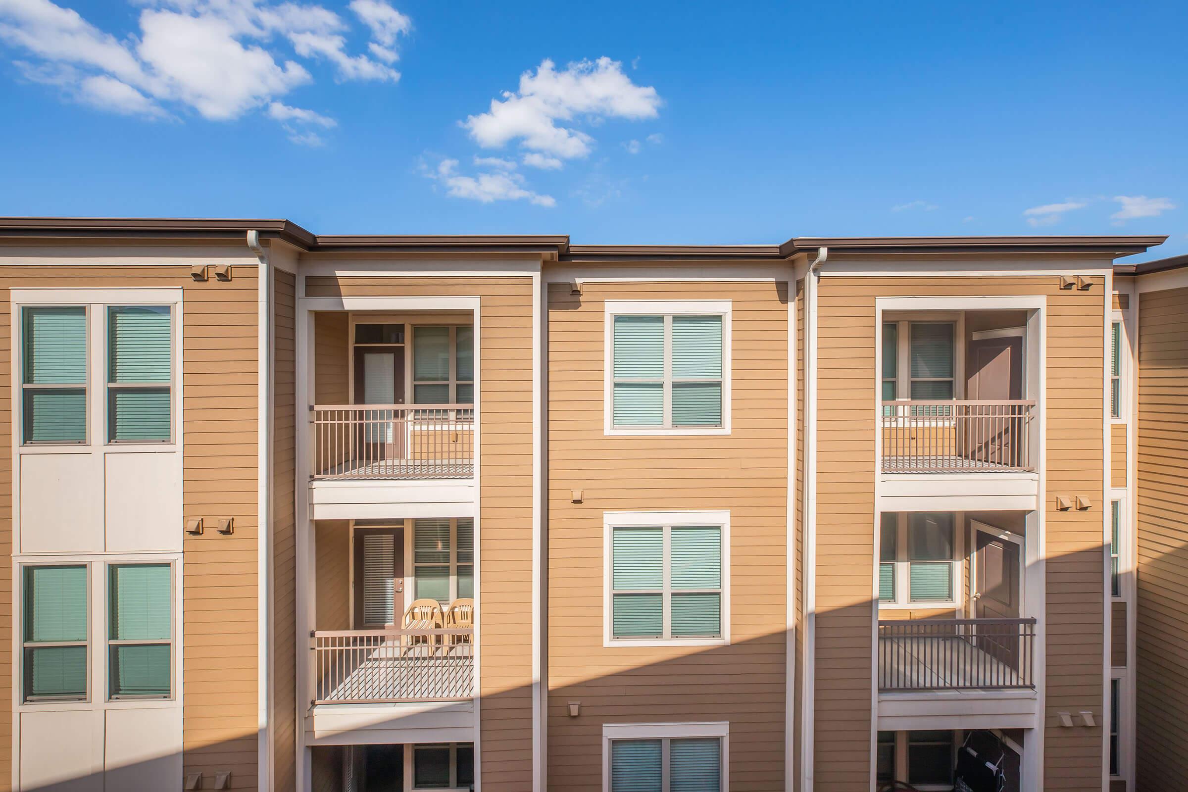 A view of an apartment building featuring three balconies, with beige siding and teal window shutters. The sky is bright blue with a few scattered clouds. Sunlight casts shadows on the building, highlighting the architectural details.