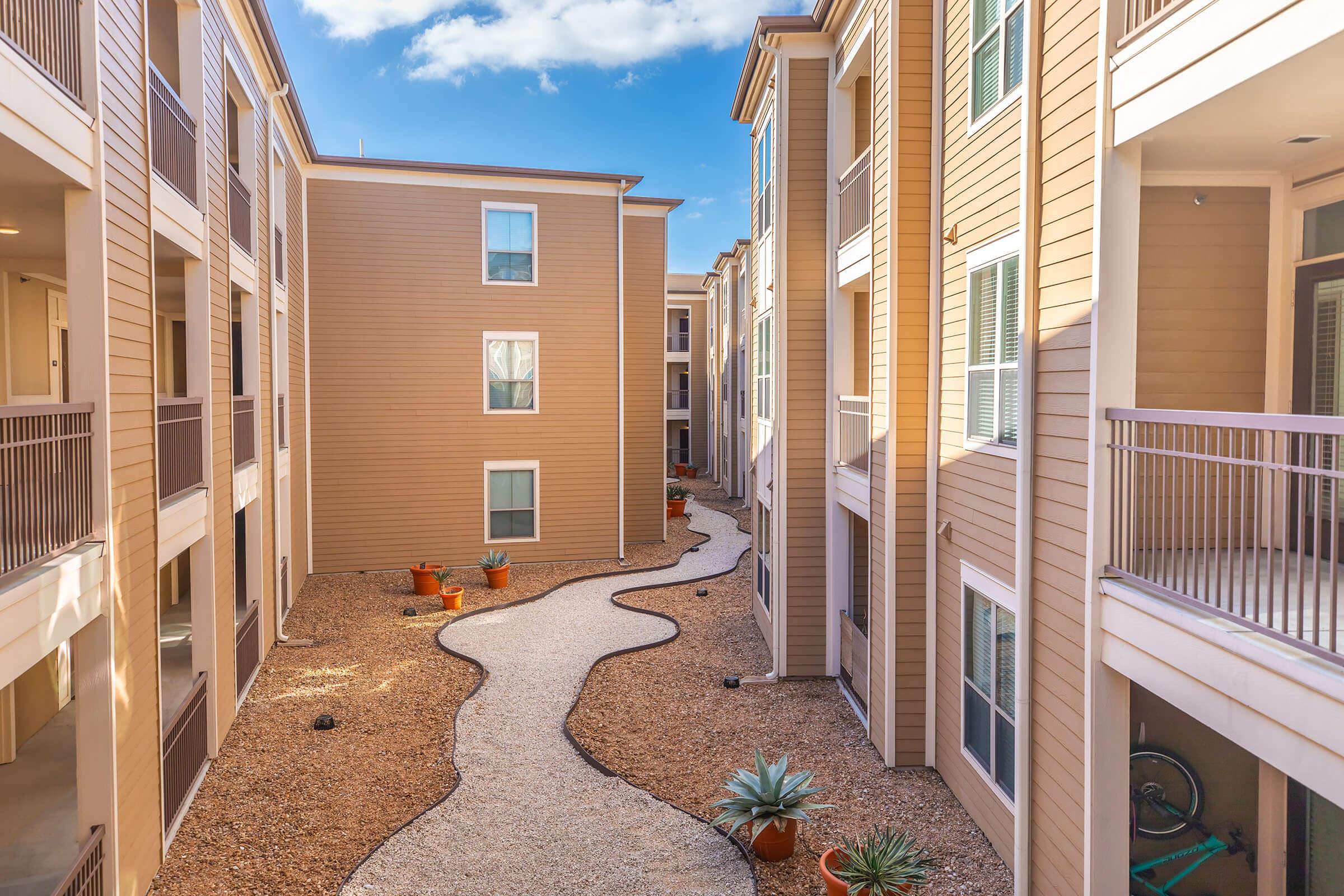 A view of a landscaped courtyard between two apartment buildings, featuring a pebbled pathway winding through the area. Potted plants are strategically placed along the sides, with balconies visible from several units. The sky is bright and blue, adding a cheerful ambiance to the scene.