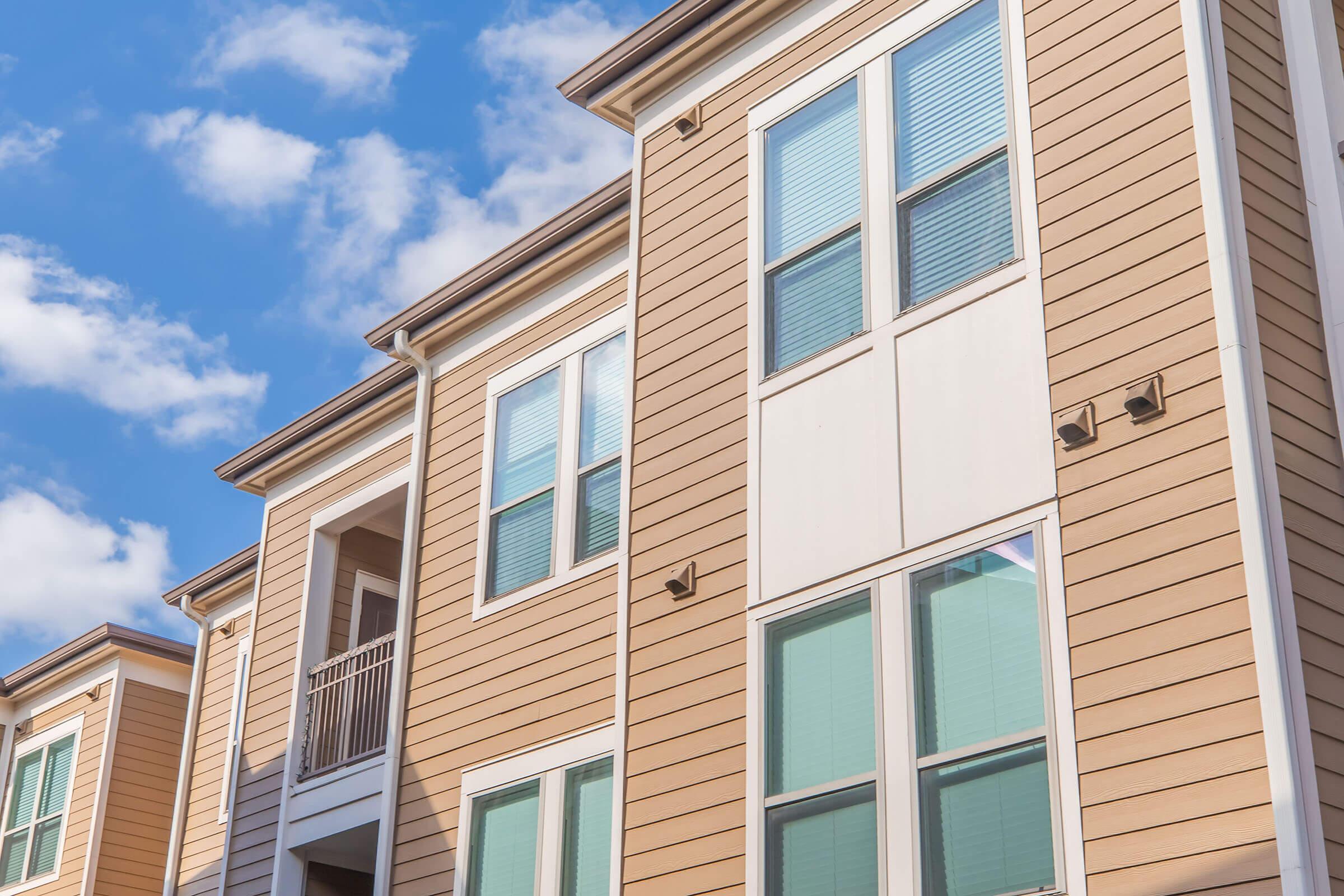 A close-up view of a modern apartment building exterior featuring light brown siding, several large windows with green reflections, and a small balcony on the second floor. The sky is bright with scattered clouds, creating a pleasant atmosphere.