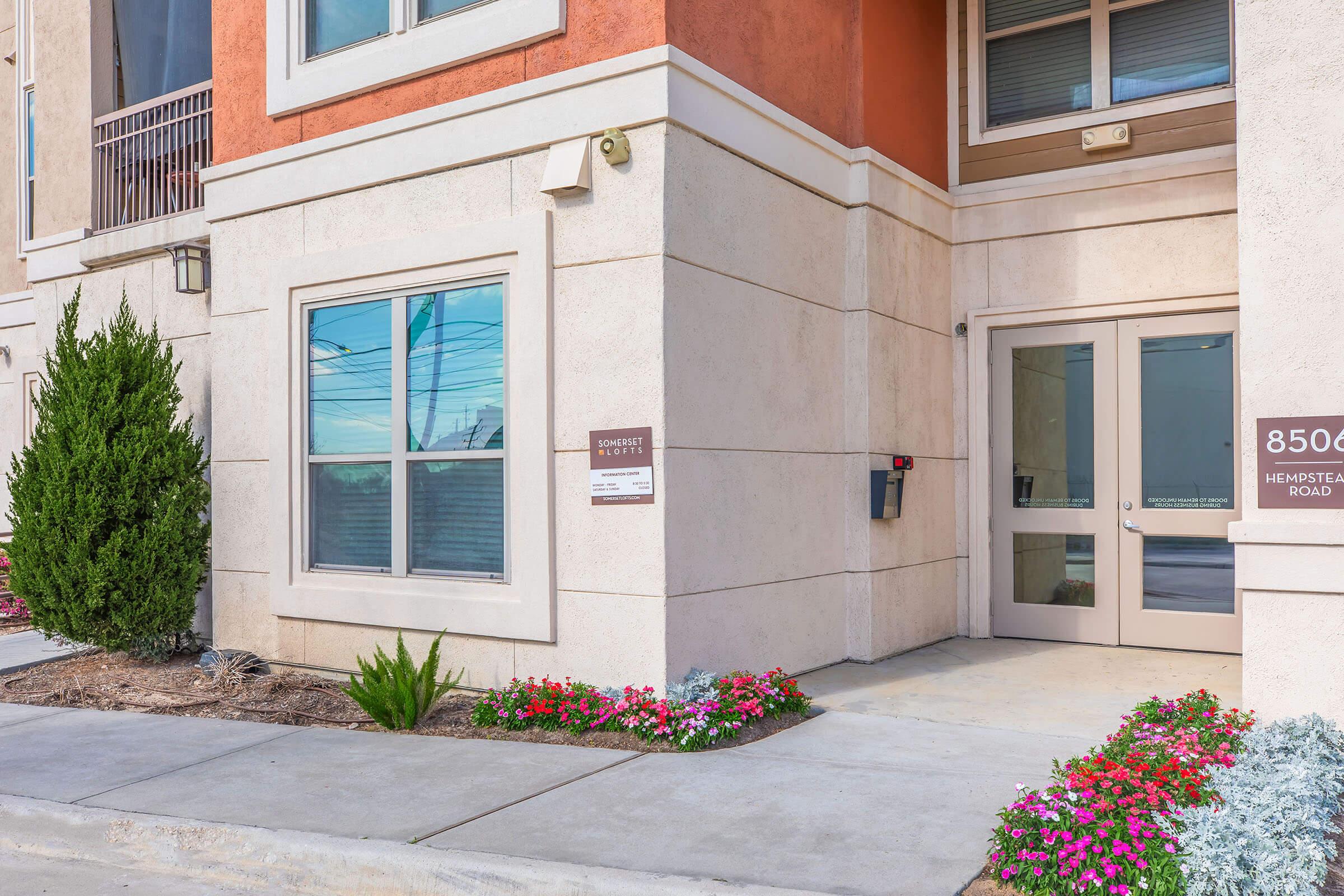 Entrance to a modern building featuring a glass double door, framed by a light-colored stone facade. Flower beds with colorful flowers and green shrubs are present along the walkways. A mailbox is mounted near the door, and the building address "8500 Hempstead Road" is displayed prominently.