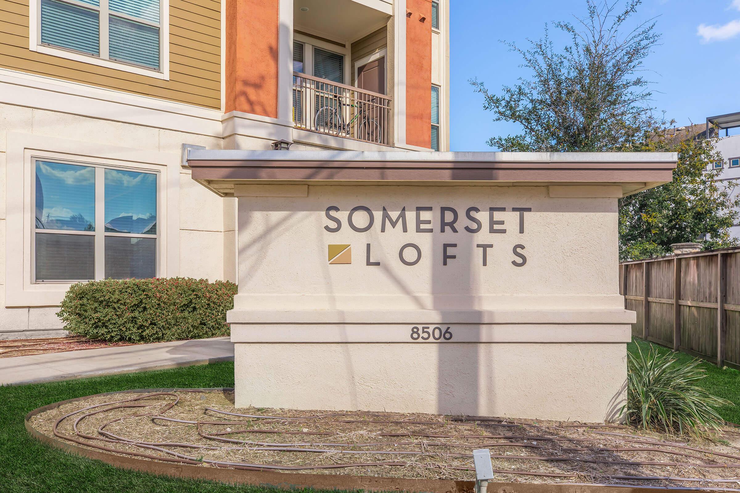 Sign for Somerset Lofts at 8506, featuring modern lettering in a clean, minimalist style against a light-colored stone base. The background includes landscaped greenery and part of a multistory building in a bright, sunny setting.