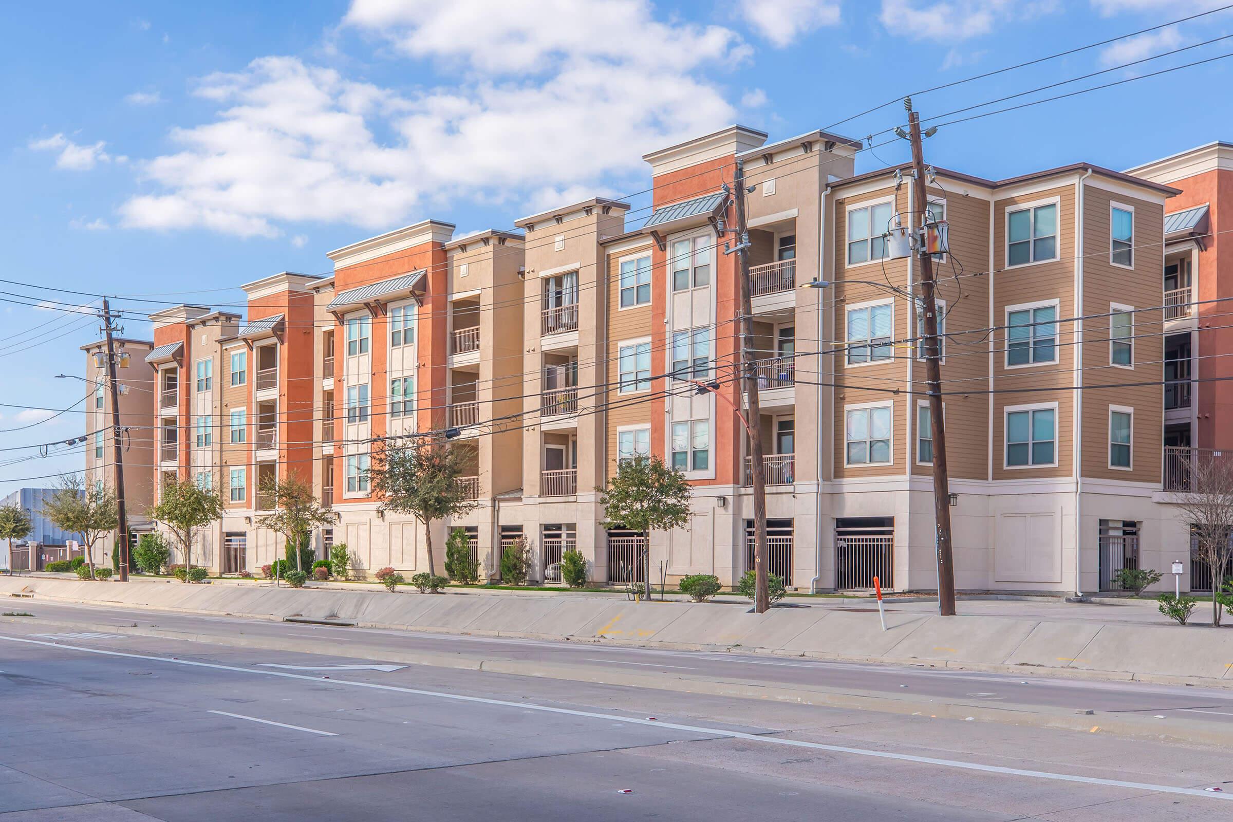 A modern multi-story apartment building with a mix of tan and red brick facades. The structure features balconies, large windows, and landscaped areas with small trees. Power lines run along the street, which is empty, and the sky is partly cloudy.