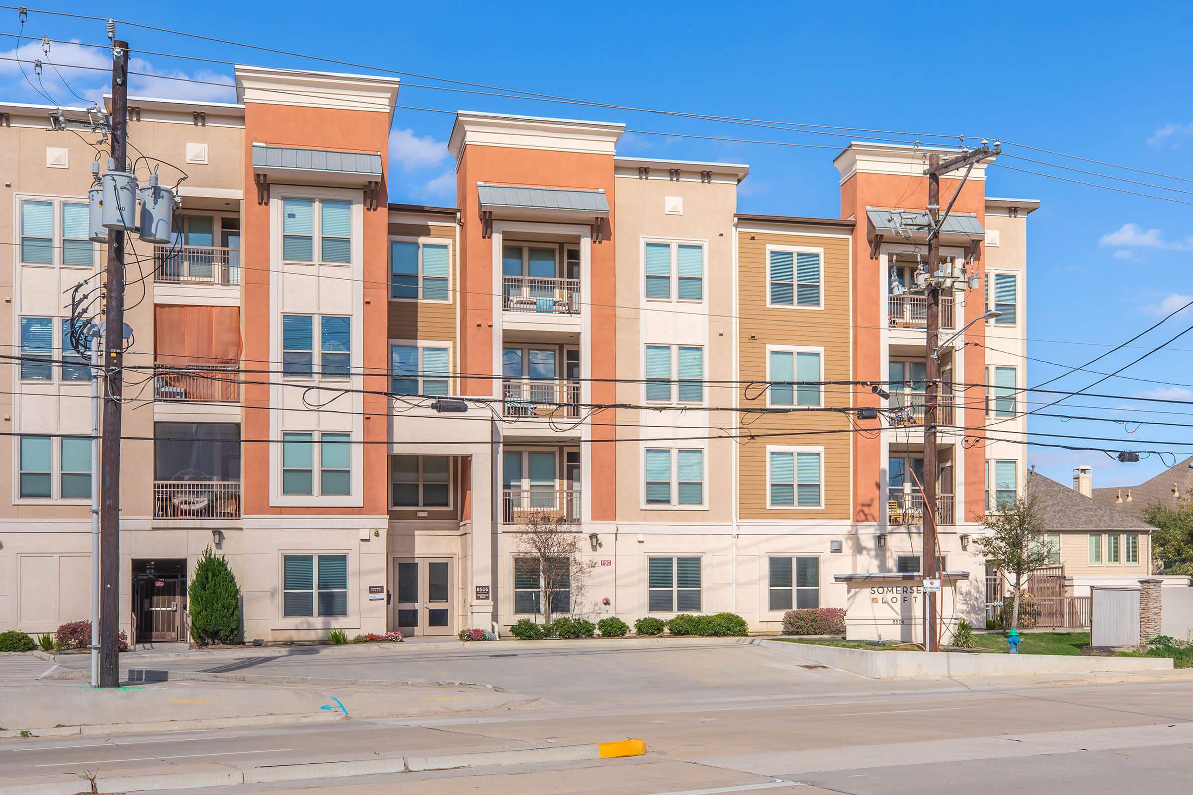 A modern apartment building with a mix of tan and orange accents. The structure features large windows and balconies. Power lines run in front of the building. The scene is set under a clear blue sky, indicating a sunny day. The surrounding area includes some greenery and residential buildings in the background.