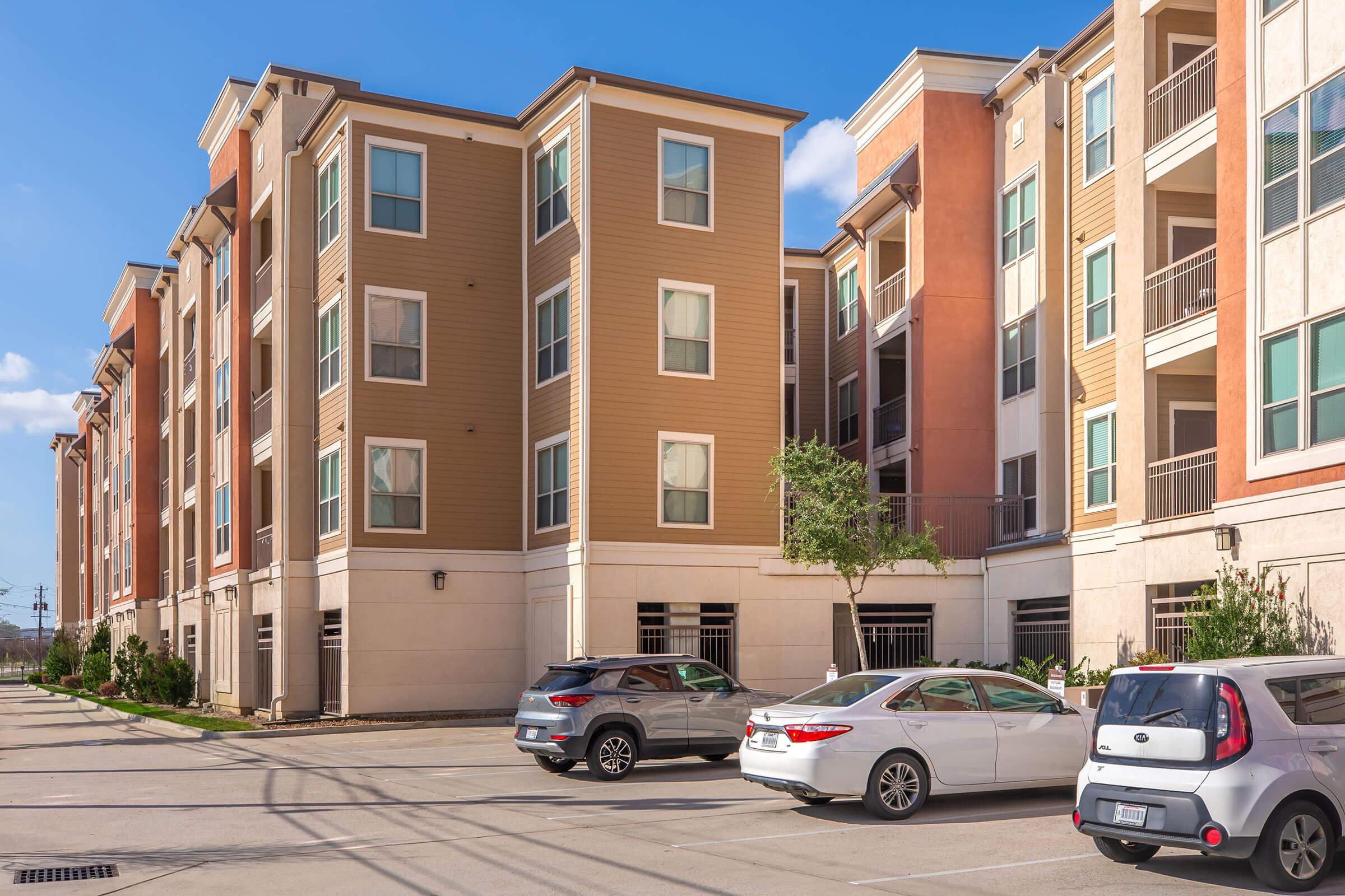 A residential apartment complex featuring multiple three to four-story buildings. The exterior includes a mix of beige and light brown siding with large windows. A parking lot in front displays several parked cars, including a compact vehicle, under a clear blue sky.