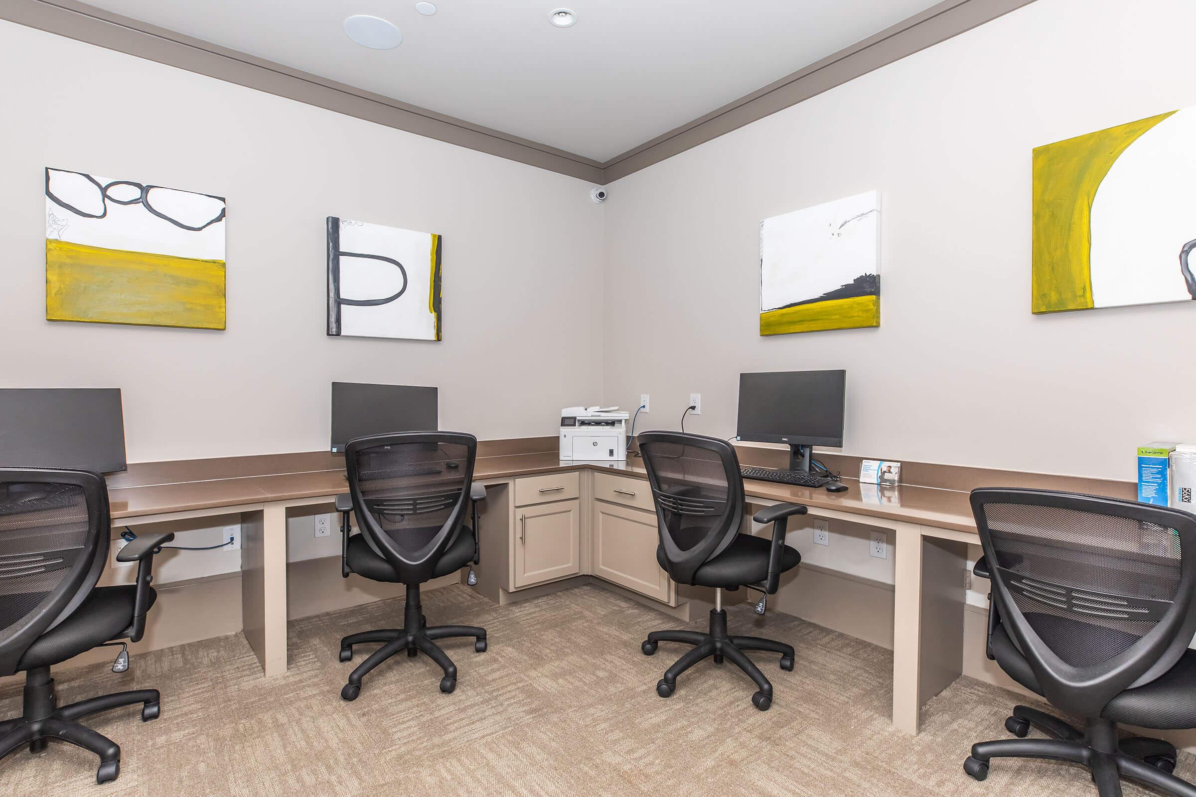 A modern office space featuring four desks with chairs, each equipped with a computer monitor. A printer is placed in the center, and the walls are adorned with abstract yellow and black artwork. The flooring is carpeted in a neutral tone, creating a professional and inviting environment.