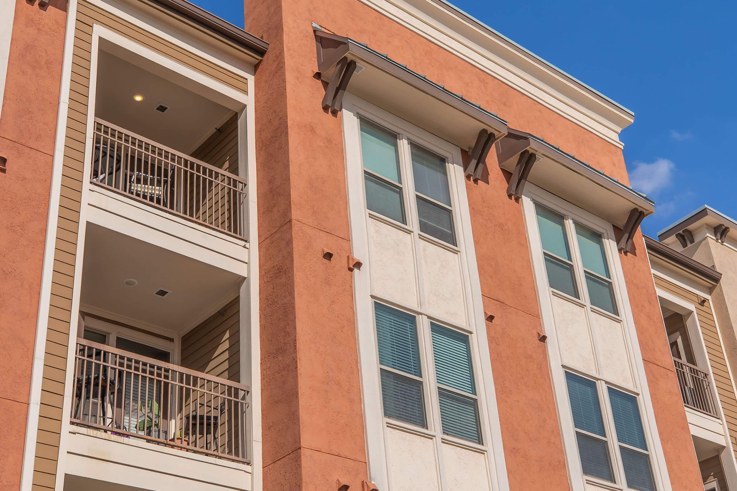 Exterior view of a modern multi-story apartment building featuring a combination of orange and beige stucco finishes. Balconies with railings are visible, along with large windows. The sky is clear with a few clouds, highlighting the architecture of the building.