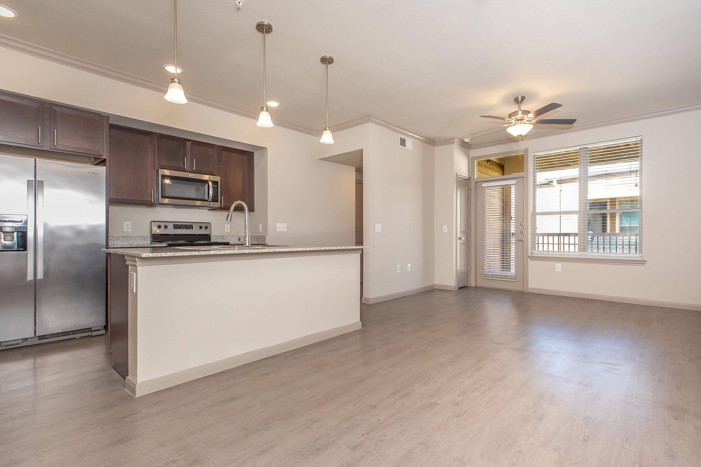 Modern open-concept kitchen and living area featuring hardwood-style flooring, stainless steel appliances, granite countertops, and pendant lighting. The space includes a ceiling fan and large windows, allowing natural light to flood in. A sliding door leads to an outdoor area.