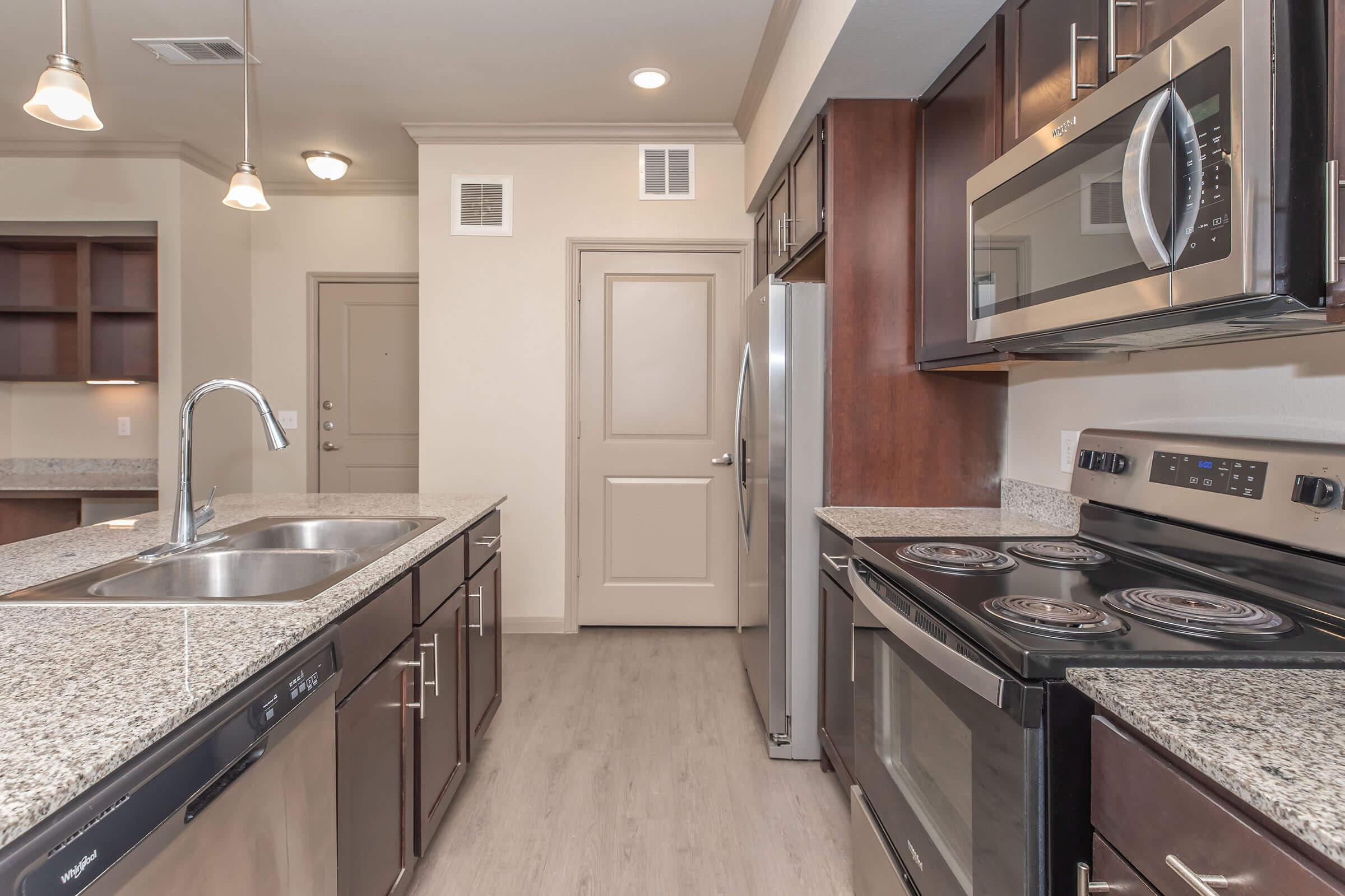 Modern kitchen featuring stainless steel appliances, including a refrigerator, oven, and microwave. The granite countertops complement dark wood cabinets. A double sink is installed in the island, and there are pendant lights overhead, creating a bright and functional cooking space.