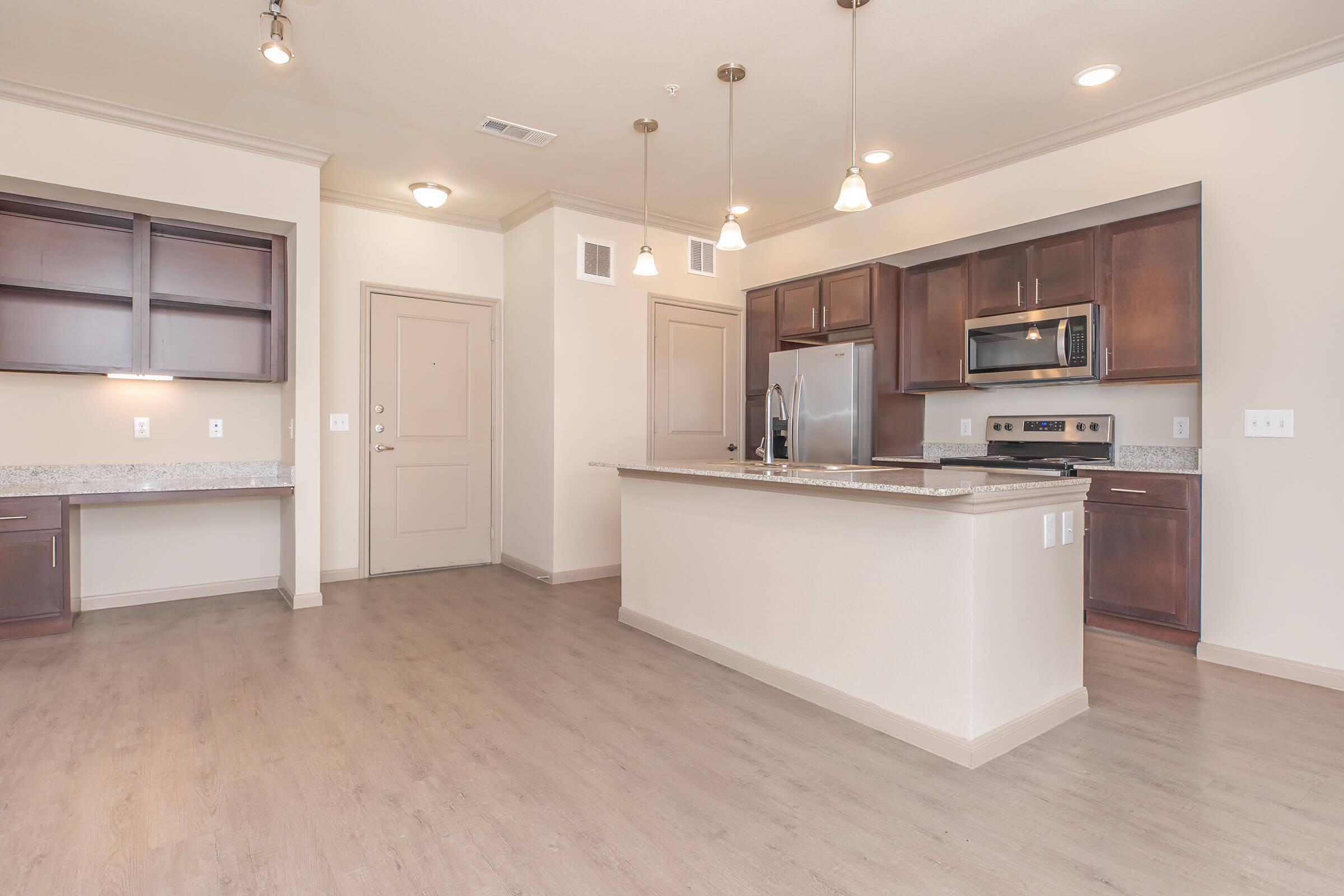 Modern kitchen and living area featuring a spacious layout, with dark wood cabinets, stainless steel appliances, and a large island with seating. The space includes hardwood-style flooring and pendant lighting, with a small nook for a desk on the left side. Neutral tones create a bright and airy atmosphere.