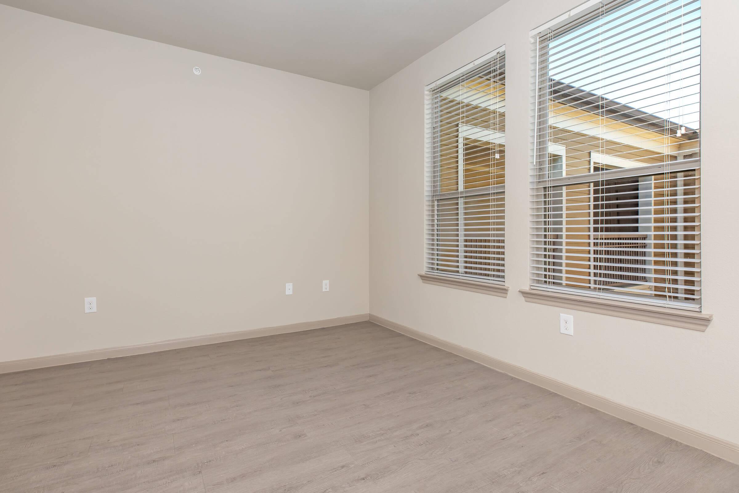 Empty room featuring light beige walls and two large windows adorned with white blinds. The floor is a light wood finish, and there are no furnishings present. Natural light streams in through the windows, creating a bright atmosphere.