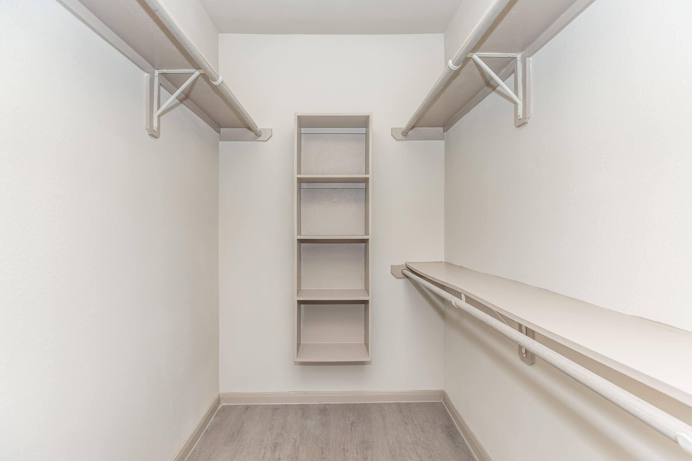 Empty walk-in closet featuring two hanging rods on either side, a central shelf unit with five cubbies, and light-colored walls. The flooring is a light wood finish, creating a clean and spacious look.