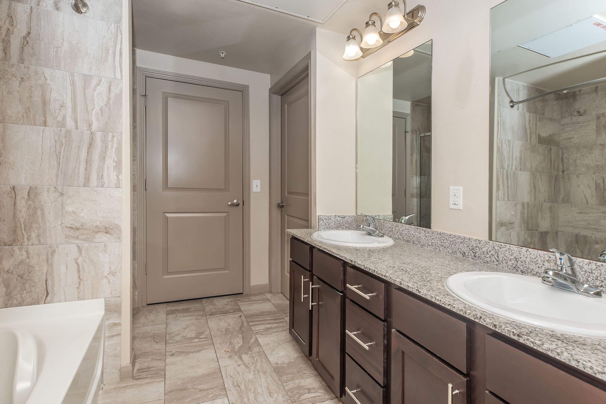 A modern bathroom featuring a double sink vanity with granite countertops, a large mirror, and a bathtub. The walls are tiled with light-colored stone, and there is a door leading to another room. Soft lighting fixtures are mounted above the mirrors, providing a warm ambiance.