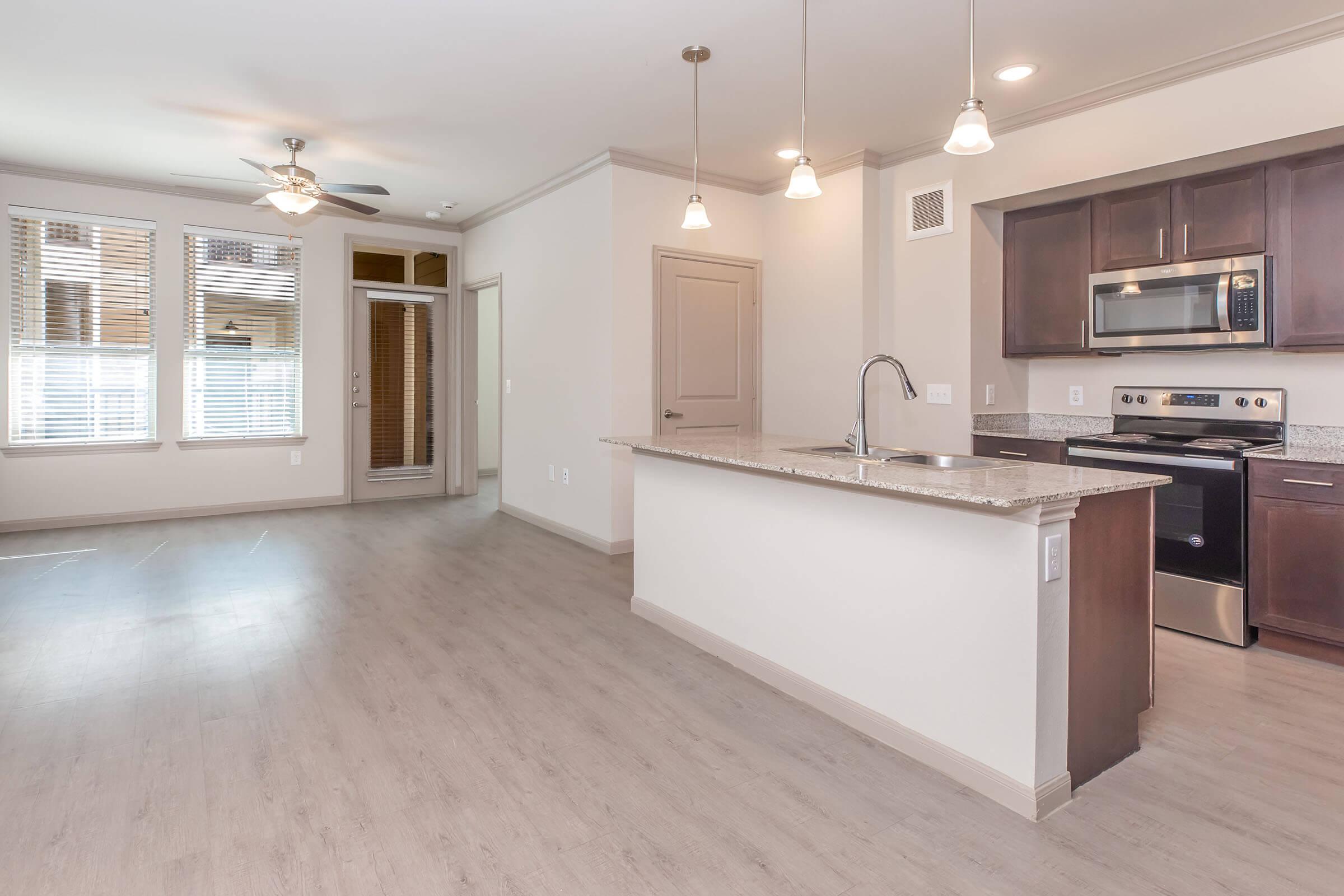 A modern apartment interior featuring an open-concept living area and kitchen. The kitchen has dark cabinetry, stainless steel appliances, and a granite countertop island with pendant lighting. Large windows provide natural light, and a ceiling fan is visible. There is a door leading to an outdoor area.