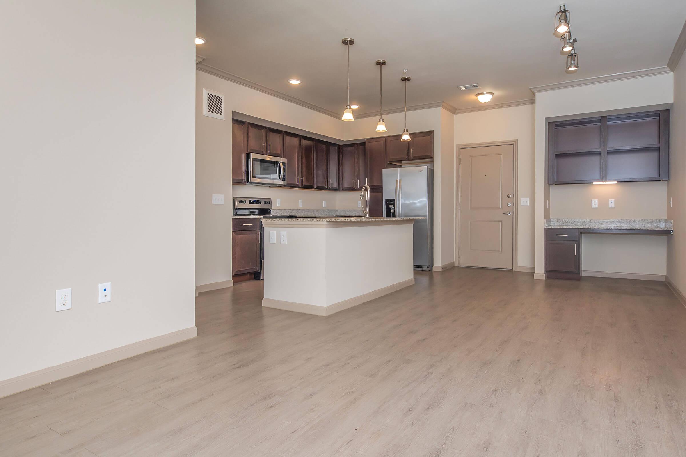 Modern kitchen and living area in a spacious apartment. Features include light-colored cabinets, stainless steel appliances, granite countertops, and pendant lighting. The open layout connects the kitchen to the living space, with wood-like flooring and beige walls creating a warm ambiance.