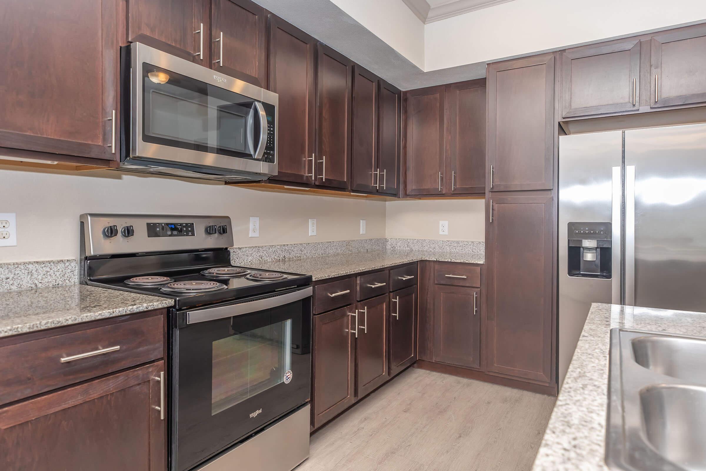 Modern kitchen featuring dark wood cabinets, stainless steel appliances including a microwave and oven, granite countertops, and a spacious layout. The kitchen includes a refrigerator with a water dispenser and a sleek design that enhances functionality and aesthetics.