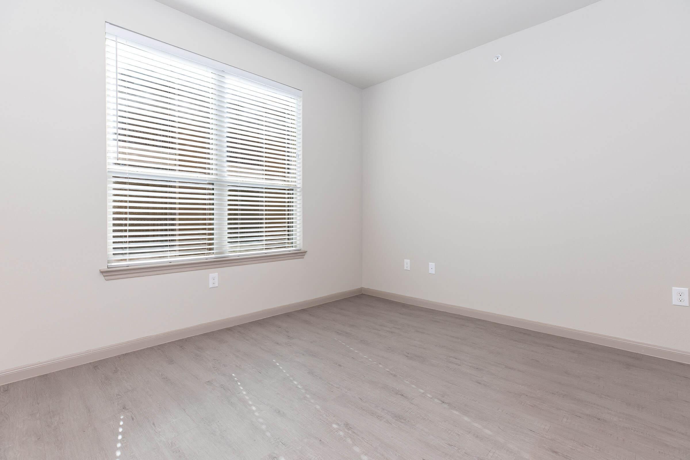 Empty room with light-colored walls and a large window featuring horizontal blinds. Natural light enters through the window, creating shadows on the floor. The flooring is a light, wood-like material, and there are no furnishings or decorations in the space.