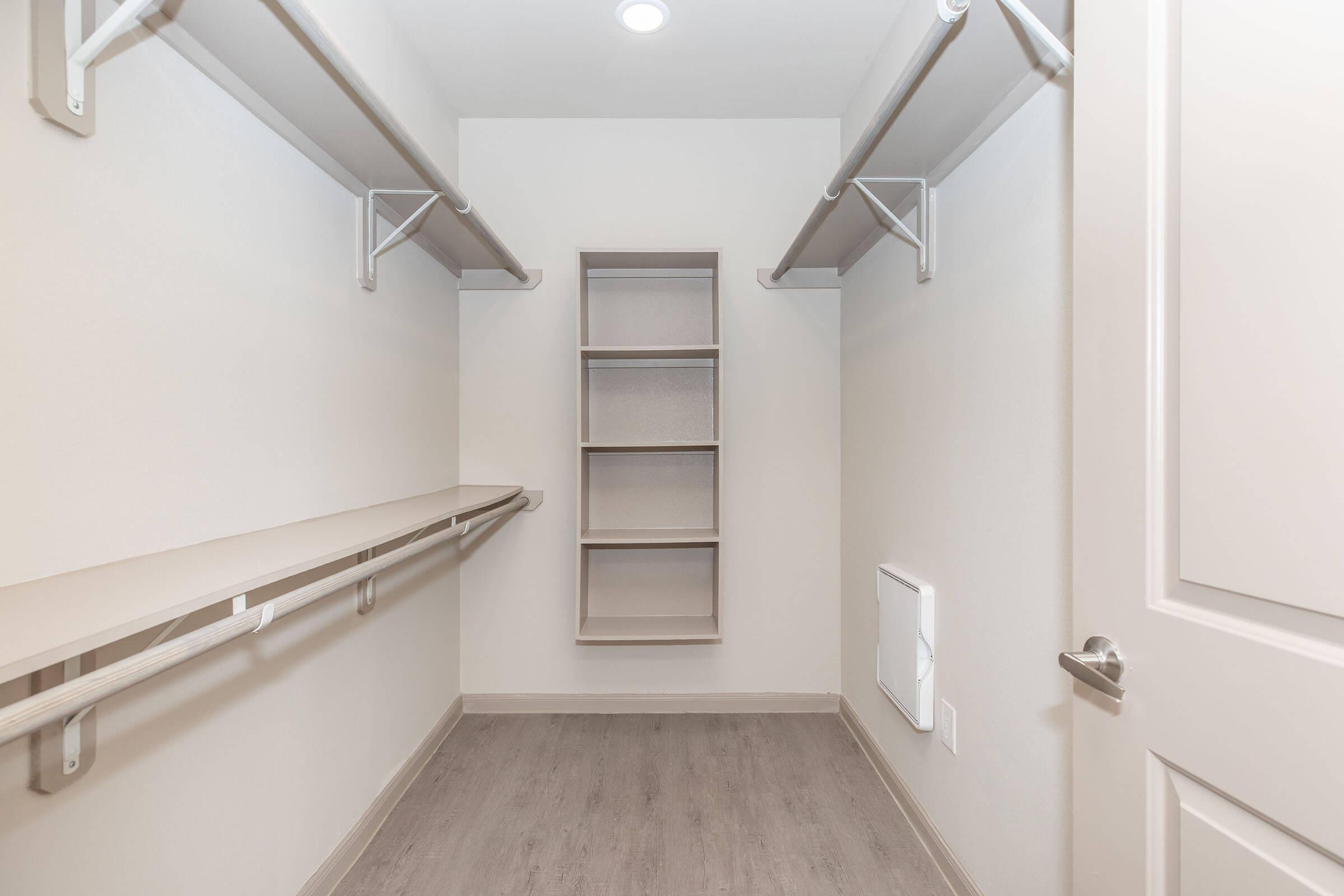 Empty closet with light-colored walls and wooden flooring. It features two horizontal clothing rods mounted on the walls and a narrow shelf unit in the center. The space is well-lit with a ceiling light, creating a bright and open feel. Doorway visible on the right side.