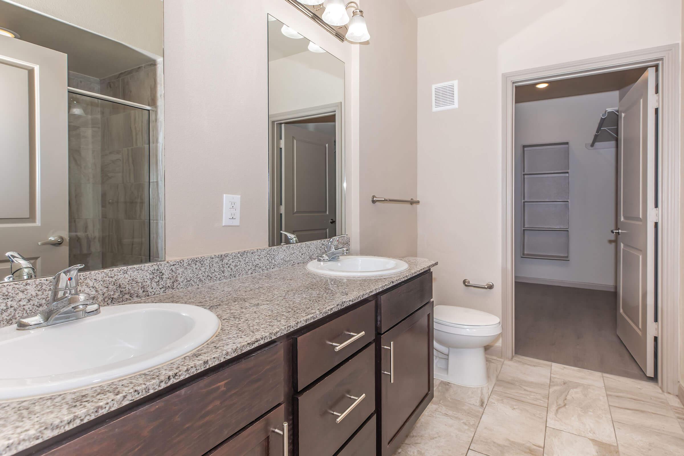 A modern bathroom featuring a double vanity with granite countertops and two sinks, a large mirror, a shower area with glass doors, and a separate toilet. The room has neutral-colored walls and tiled flooring, with an adjacent closet space visible through an open door.