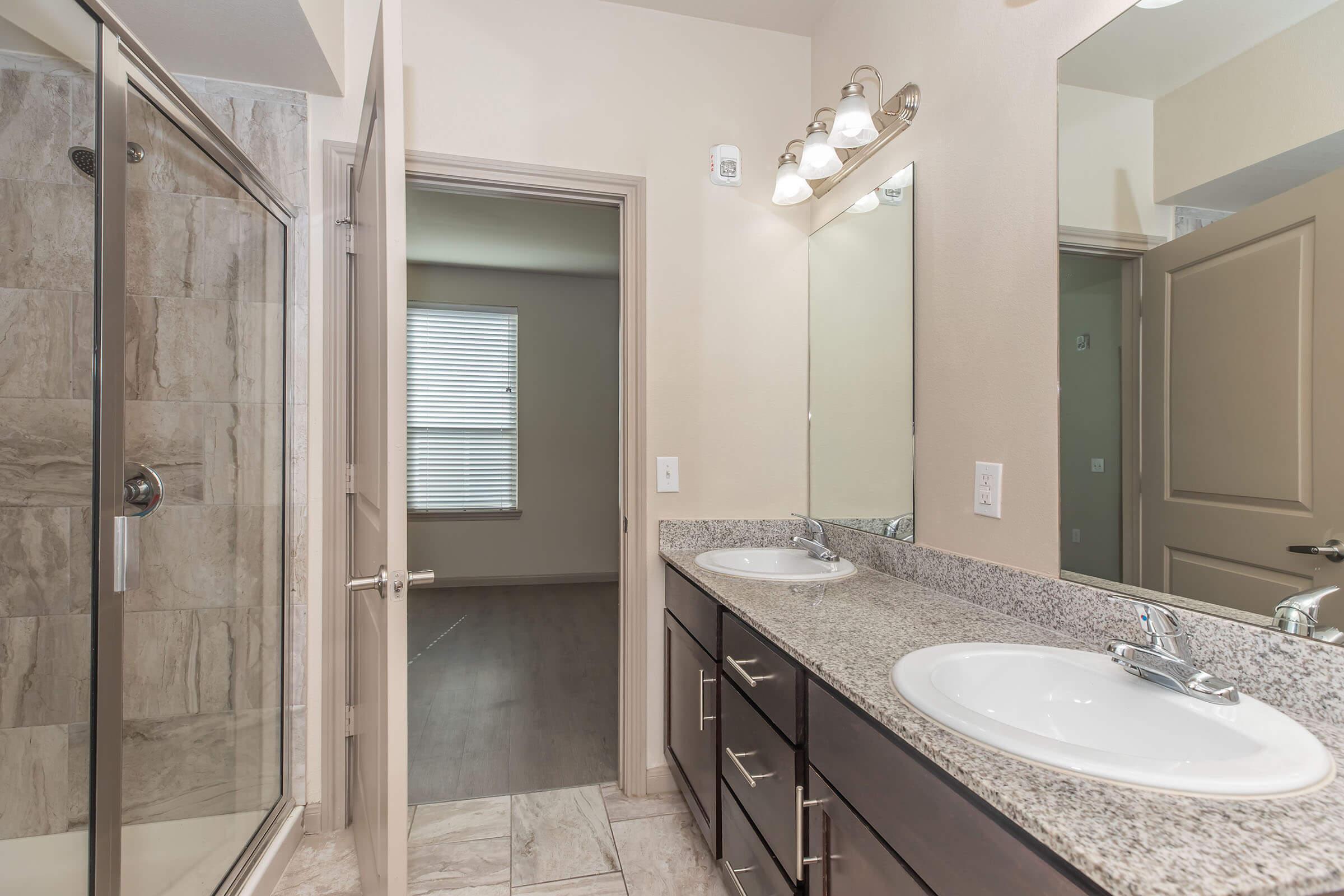 A modern bathroom featuring a double vanity with two sinks, dark wood cabinets, and a large mirror. There is a glass shower enclosure with tiled walls and flooring, and a door leading to another room with natural light coming through a window.