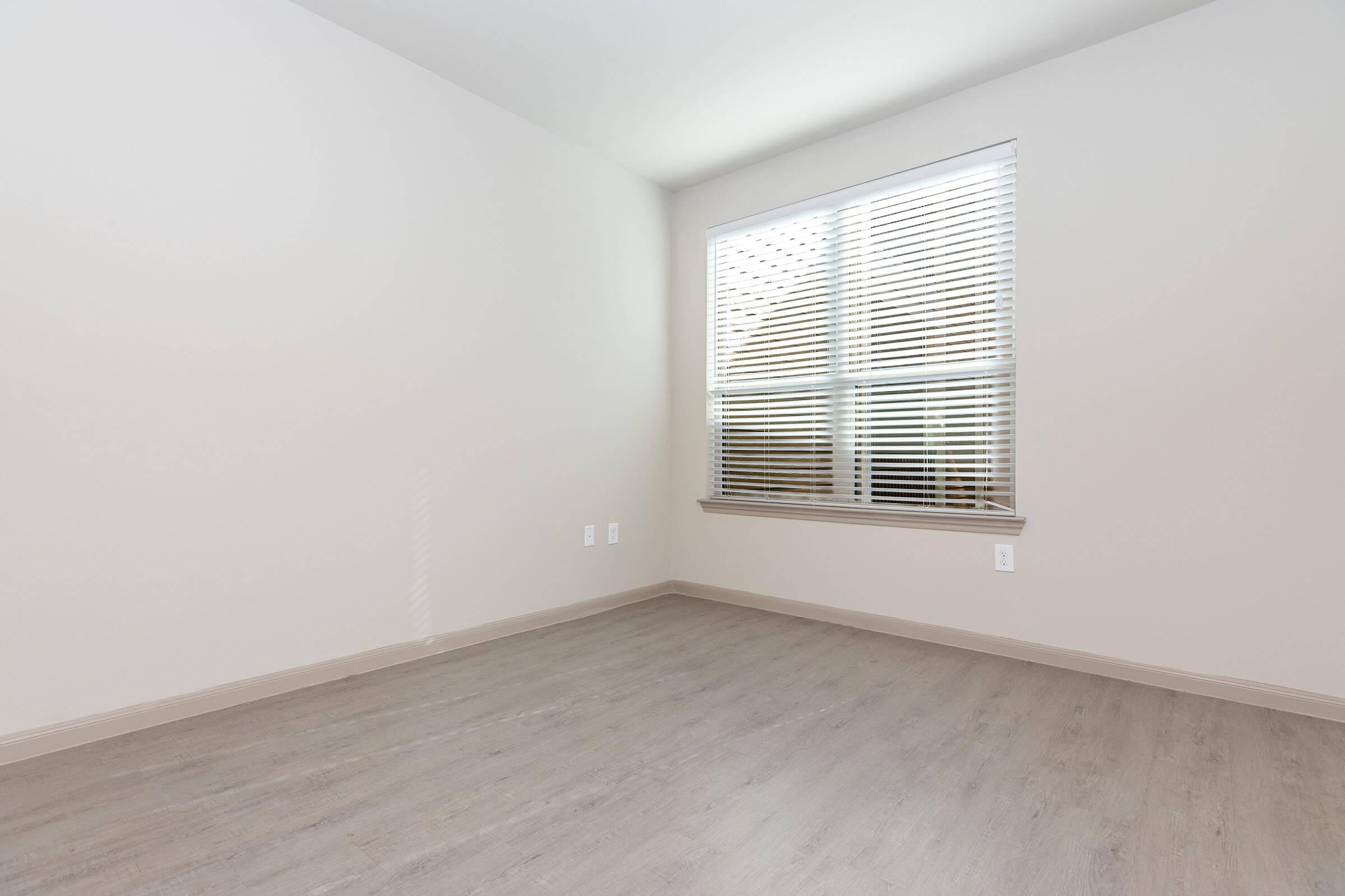 Empty room with light-colored walls and a window dressed with horizontal blinds. The floor is a light wood finish. The space is well-lit, creating a bright atmosphere.