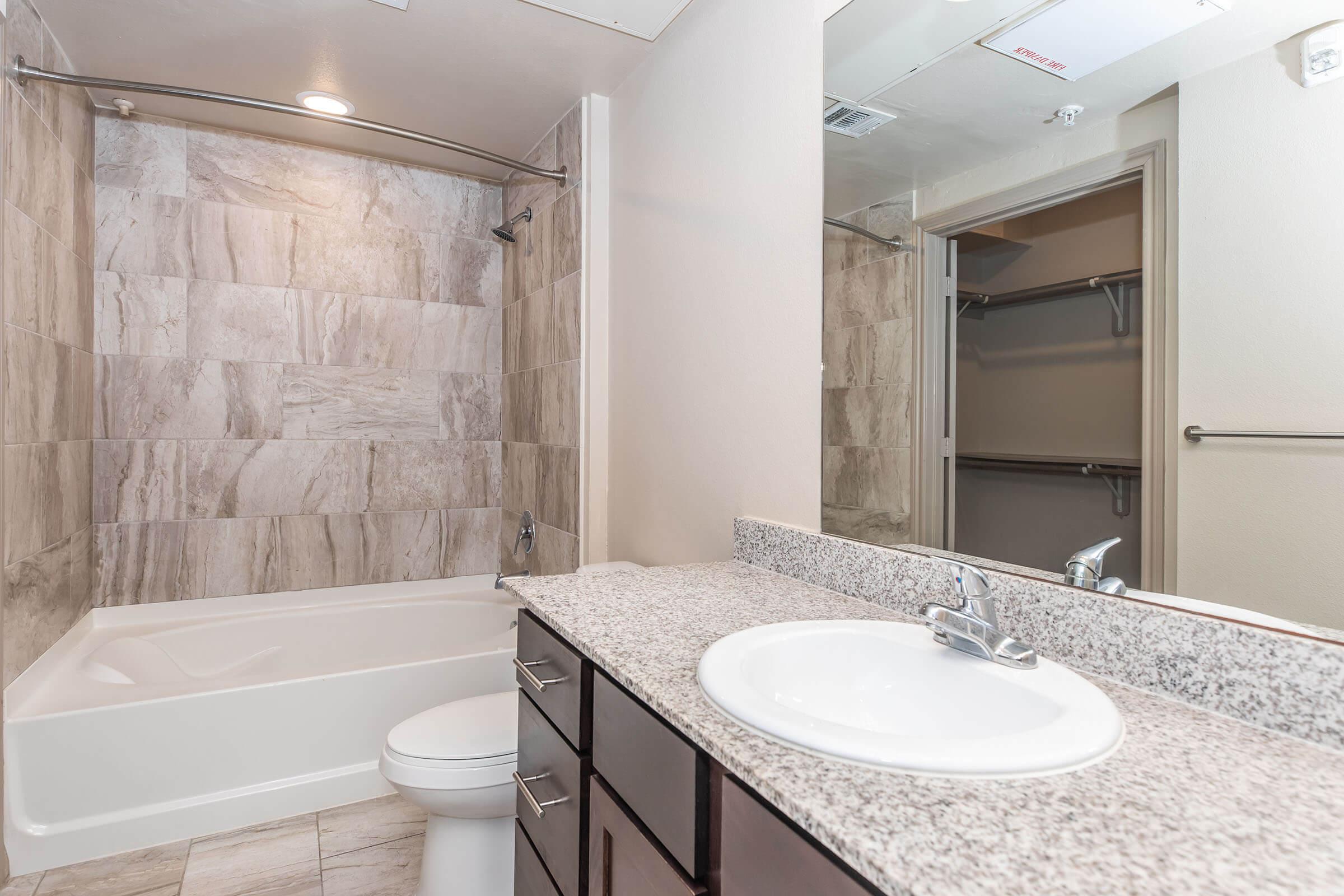 A modern bathroom featuring a bathtub with a shower, beige and gray tiled walls, a granite countertop with a sink, and dark wood cabinetry. A large mirror reflects the space, and a closet can be seen in the background. Bright lighting enhances the clean, stylish design.