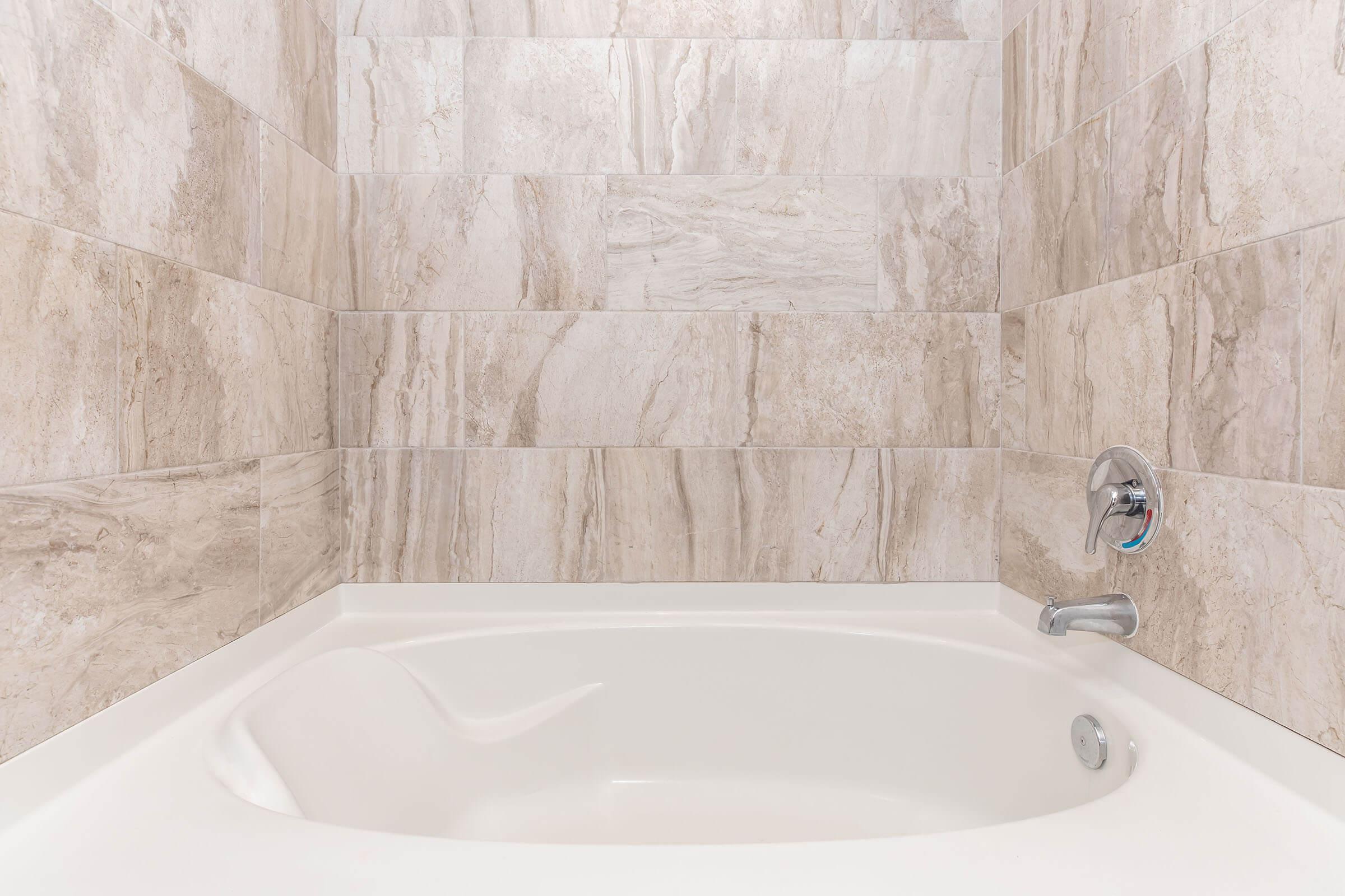 A porcelain bathtub set against tiled walls featuring a light beige and gray marbled design. The tub has a chrome faucet and a temperature control handle, creating a modern bathroom aesthetic. The clean and minimalist look emphasizes a serene bathing space.