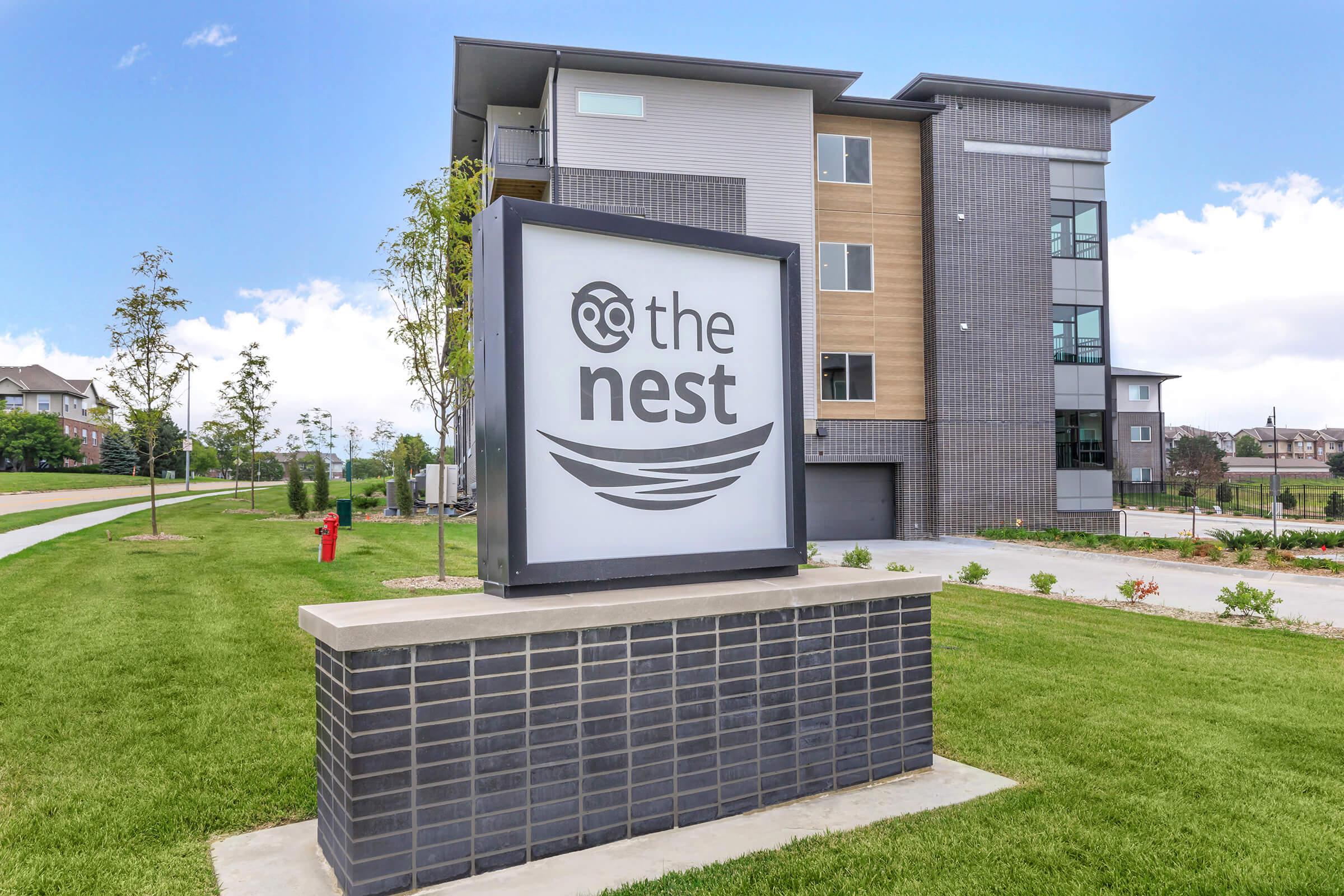 A sign for "the nest" featuring a modern design, located in front of a contemporary multi-story building. The building has a combination of gray and beige exterior materials, and the surrounding area includes well-manicured grass and trees.