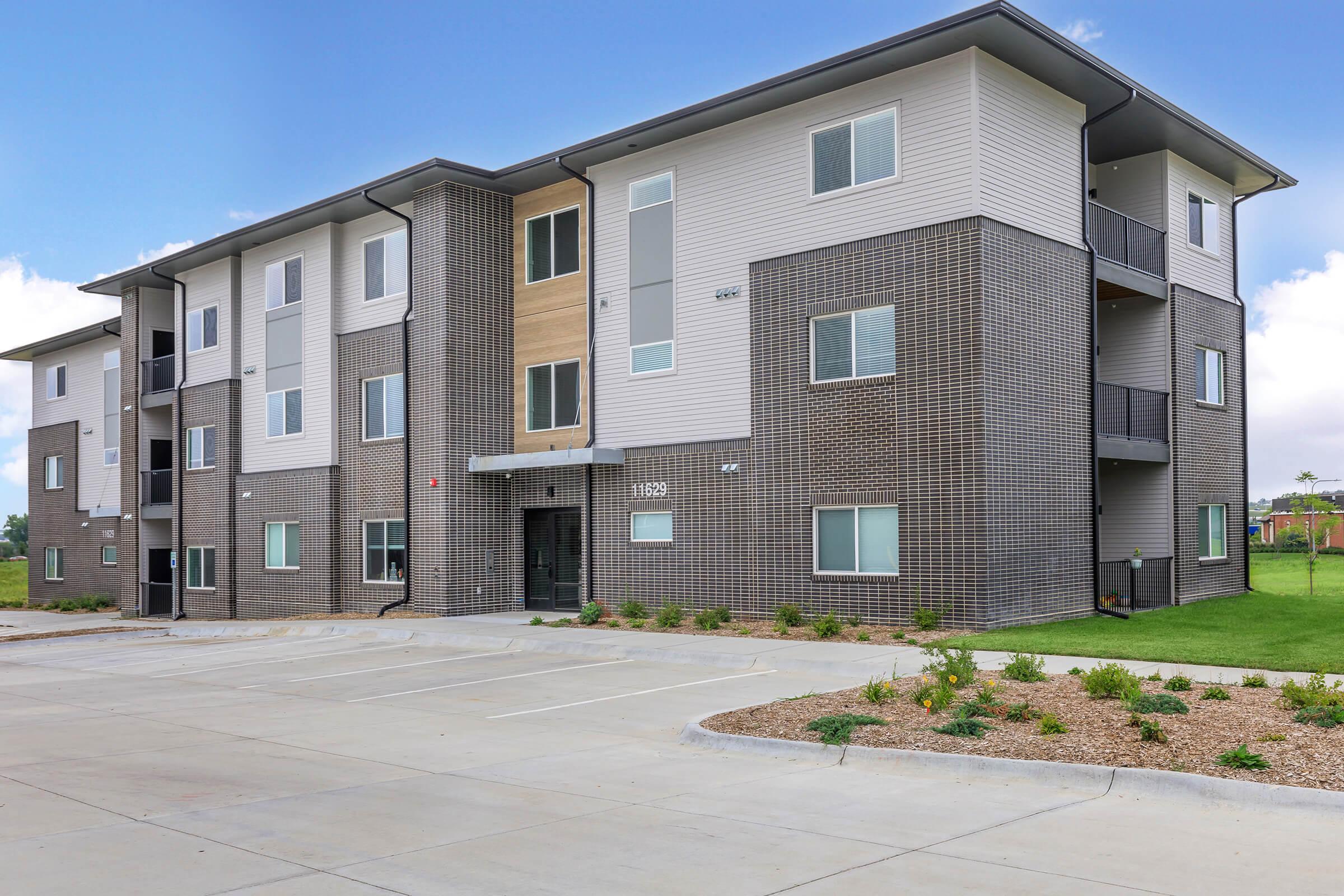 A modern three-story apartment building featuring a mix of brick and siding exterior, with several windows and a main entrance. Surrounding the building is a parking area and landscaped patches, indicating a well-maintained residential complex. The sky is clear with minimal clouds.