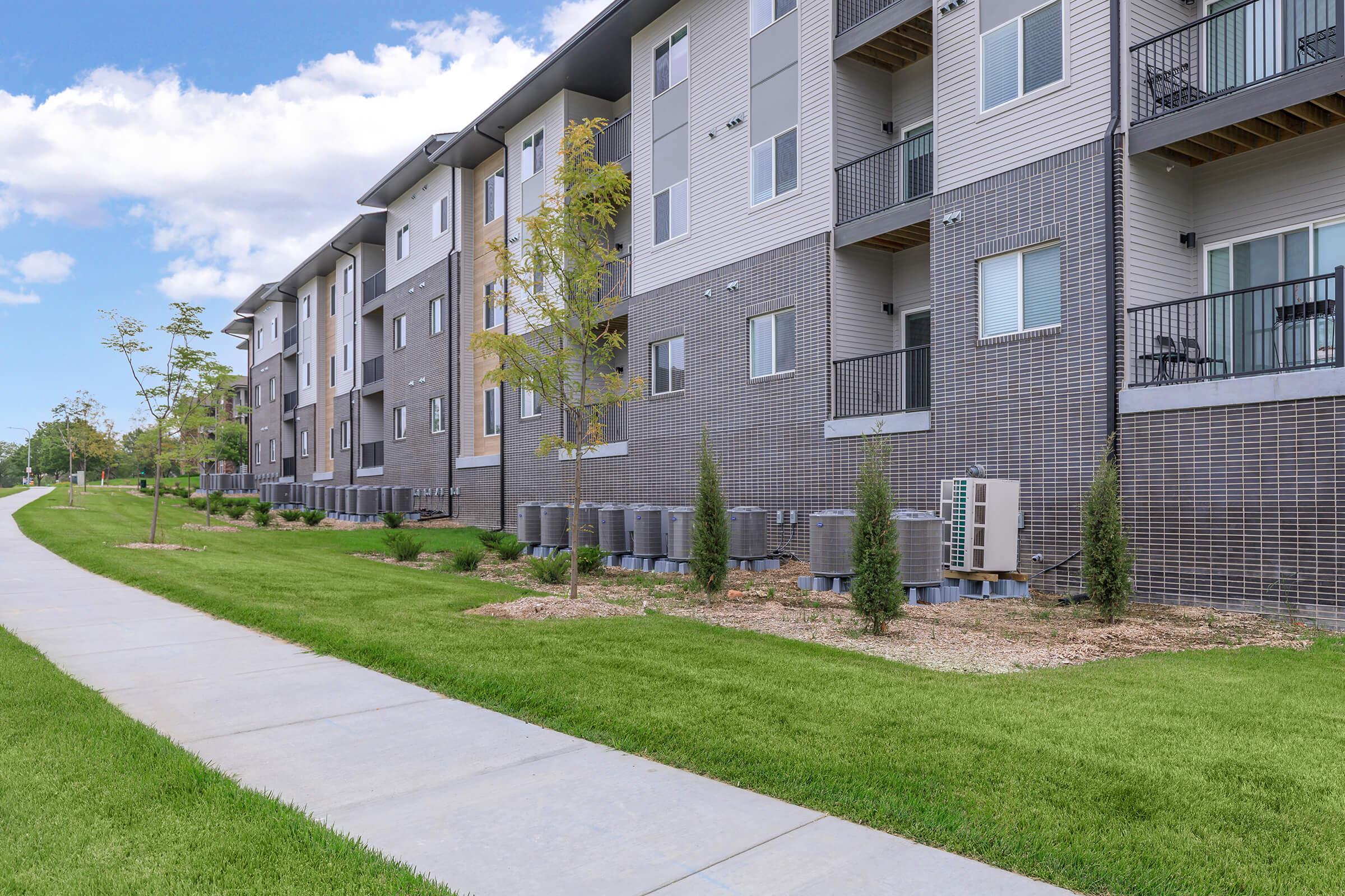Modern apartment building with several units featuring balconies, situated alongside a paved walkway. Well-maintained green lawn and landscaping with small trees and shrubs add to the aesthetic. Air conditioning units are visible along the building's exterior. Bright blue sky with a few clouds in the background.