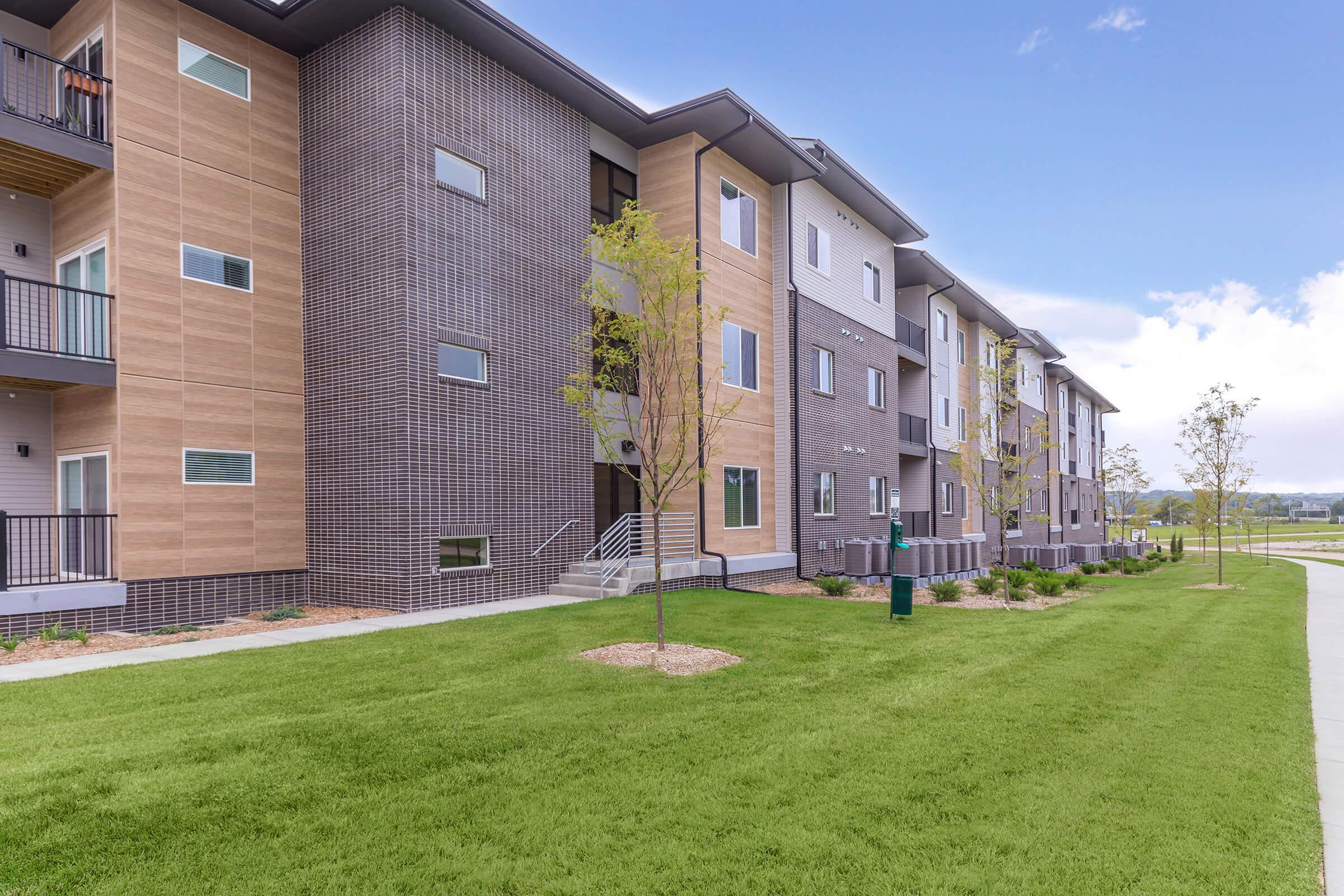 Modern multi-story apartment building featuring a blend of light wood and dark brick exteriors. The structure has multiple balconies, large windows, and is surrounded by green lawns and young trees. A paved walkway runs alongside, enhancing the inviting atmosphere. Clear blue sky in the background.