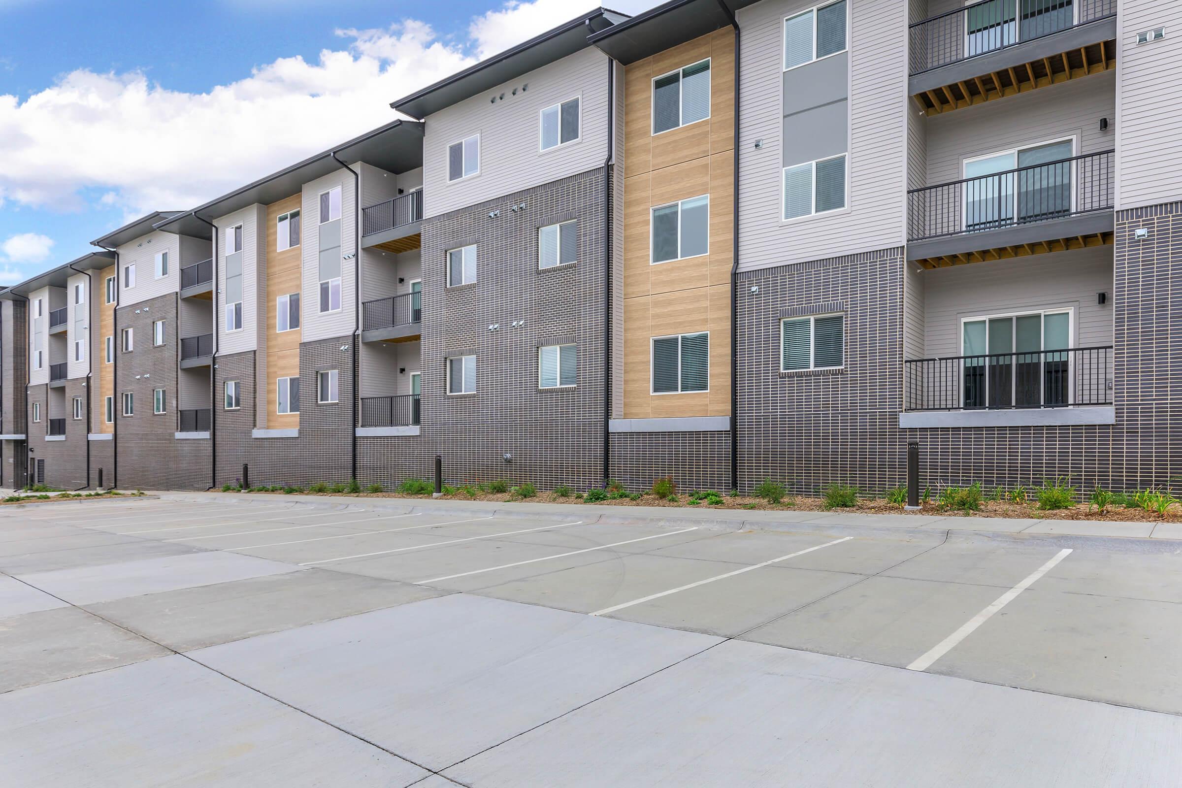 A modern apartment building featuring multiple floors with a mix of light and dark siding. Windows are prominently displayed on each unit, and there is a spacious, empty parking lot in the foreground with several marked parking spaces. The sky is partly cloudy, adding depth to the scene.