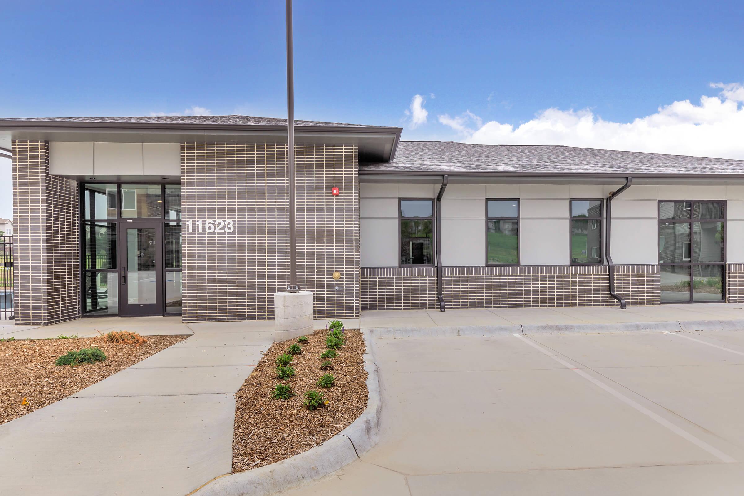 A modern building with a brick facade, featuring large windows and a glass entrance. The address "11623" is displayed on the wall. The surrounding area has a well-maintained landscape, including mulch and small green plants, with a paved parking area in front. The sky above is clear with a few clouds.