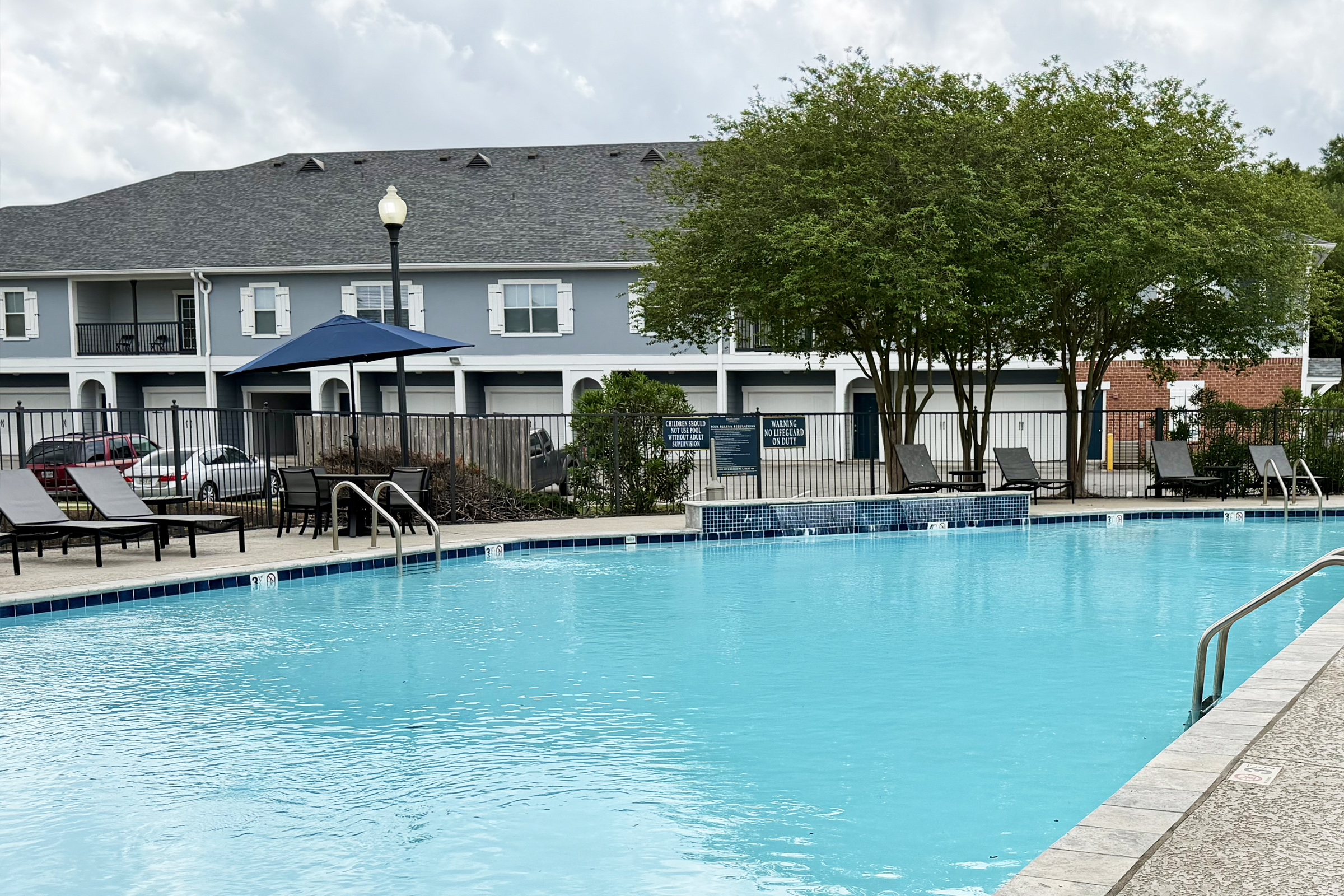 A sparkling blue swimming pool surrounded by lounge chairs and an umbrella. In the background, there are residential buildings with balconies and greenery, creating a relaxing outdoor leisure area. The sky is partly cloudy, adding to the tranquil atmosphere.